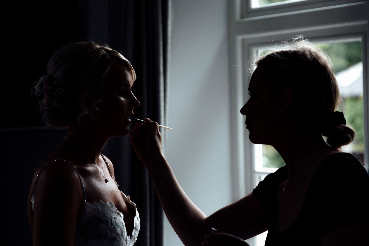 Silhouetted figures share intimate moments near a window during wedding preparations.