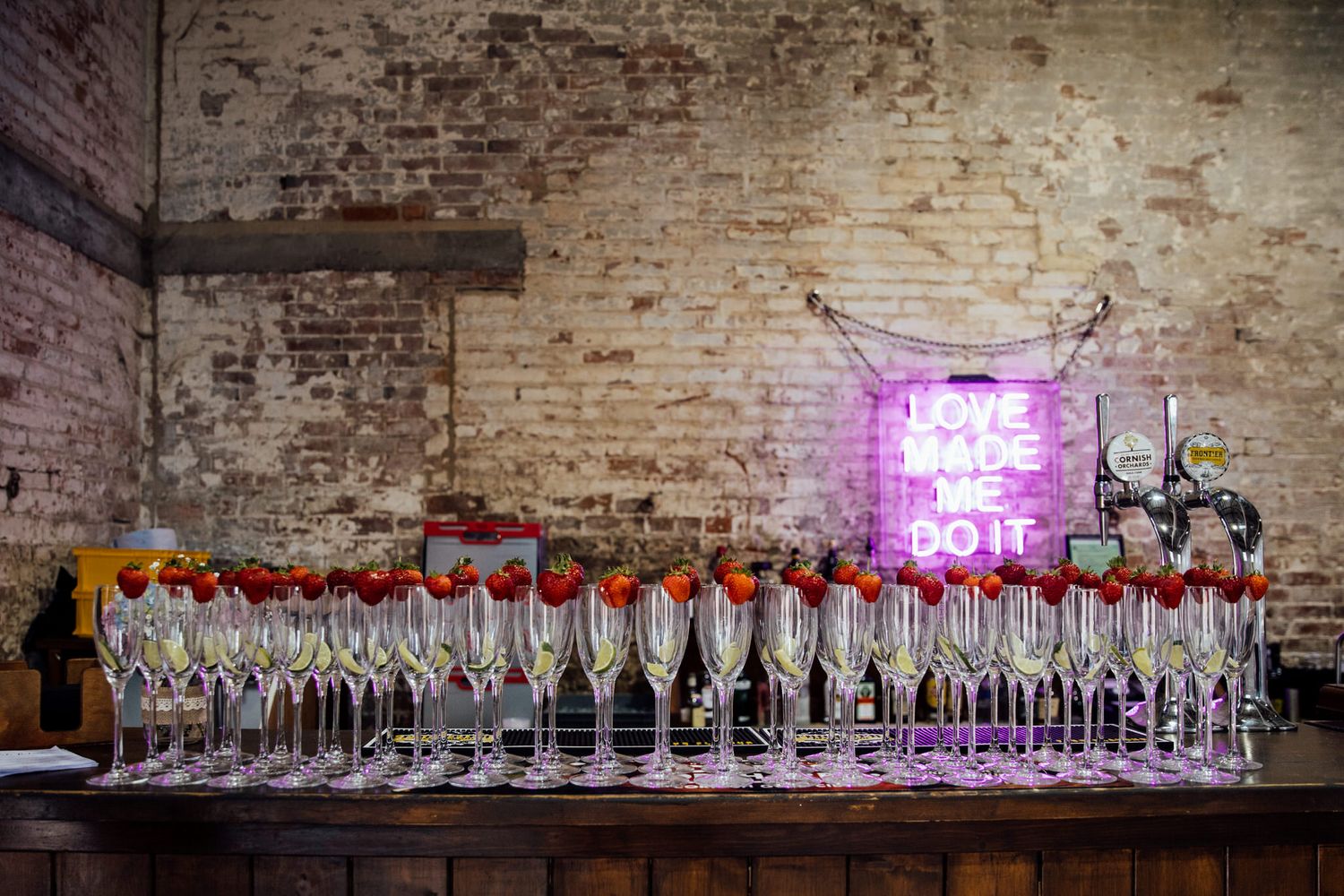 Long table display of wine bottles and glasses against exposed brick wall with pink lighting.