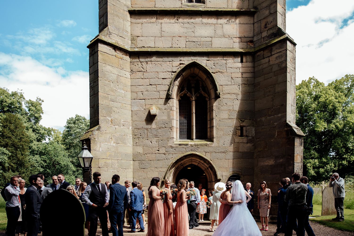Wedding ceremony guests gathered outside a stone church on a sunny summer day.