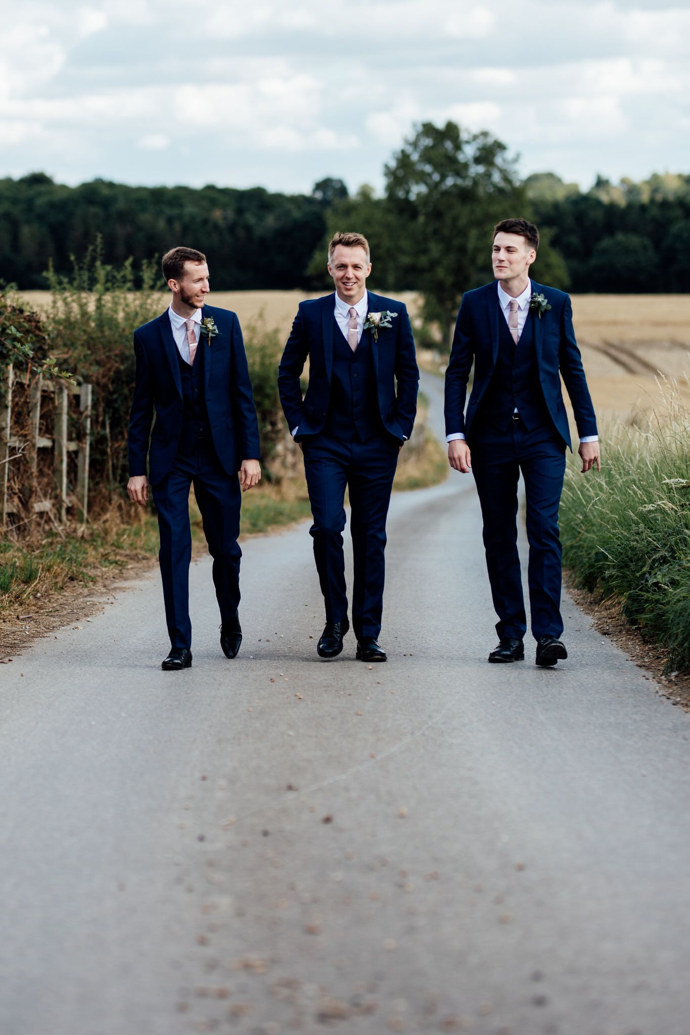 Groomsmen in navy suits walk along a country path surrounded by fields.