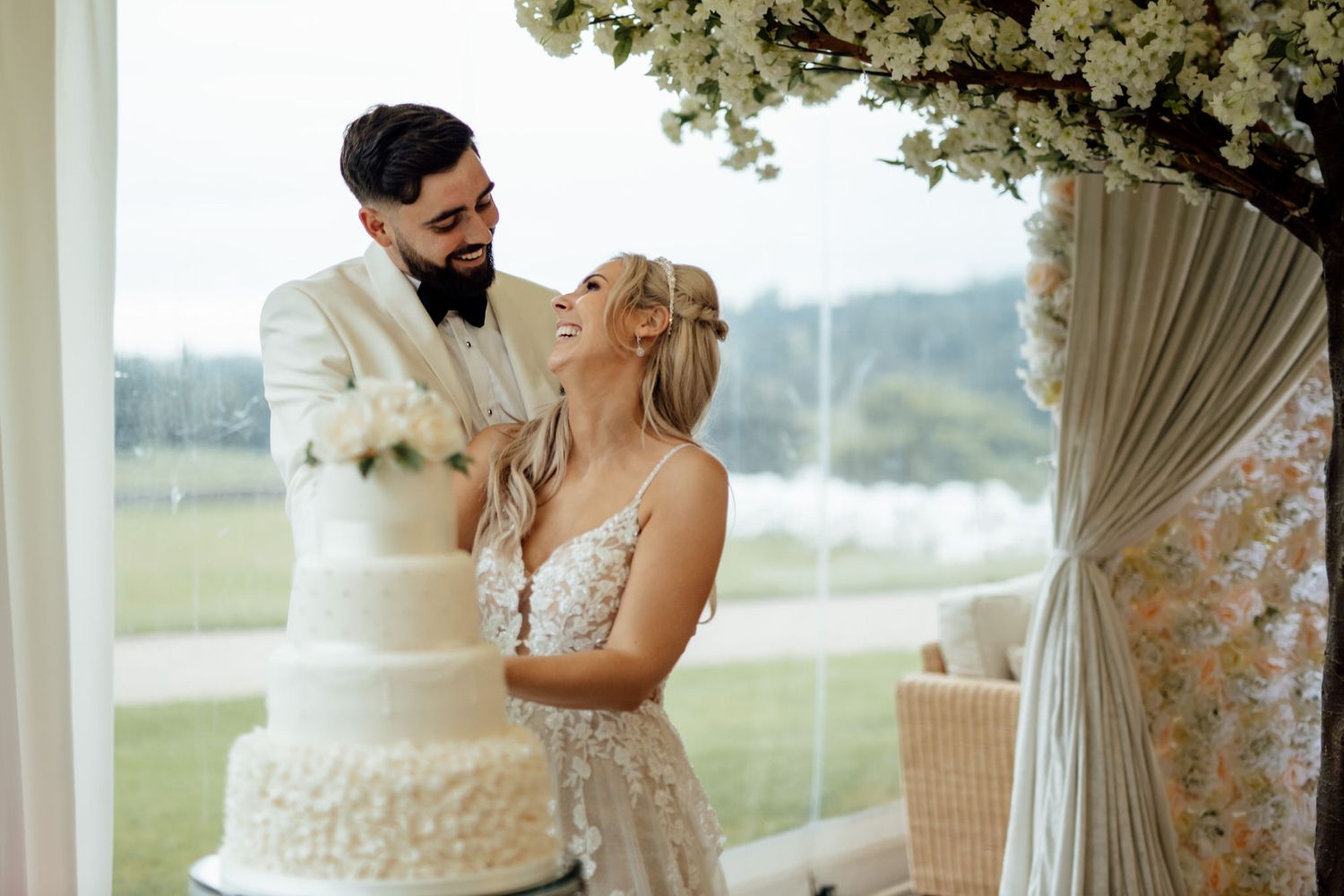 Elegant white tiered wedding cake displayed by window with floral decorations.