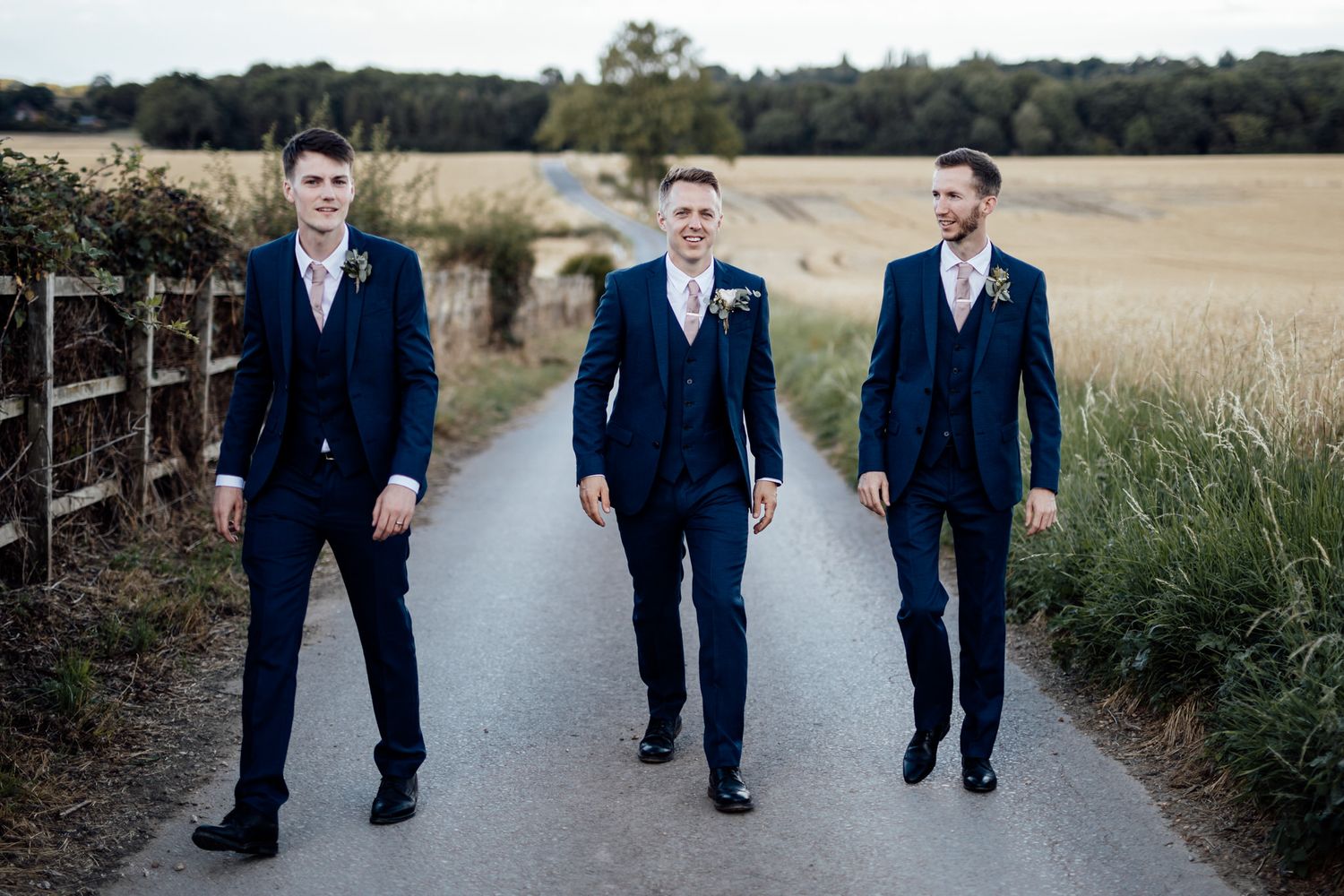Three groomsmen in navy suits walk confidently down rural path near wooden fence.