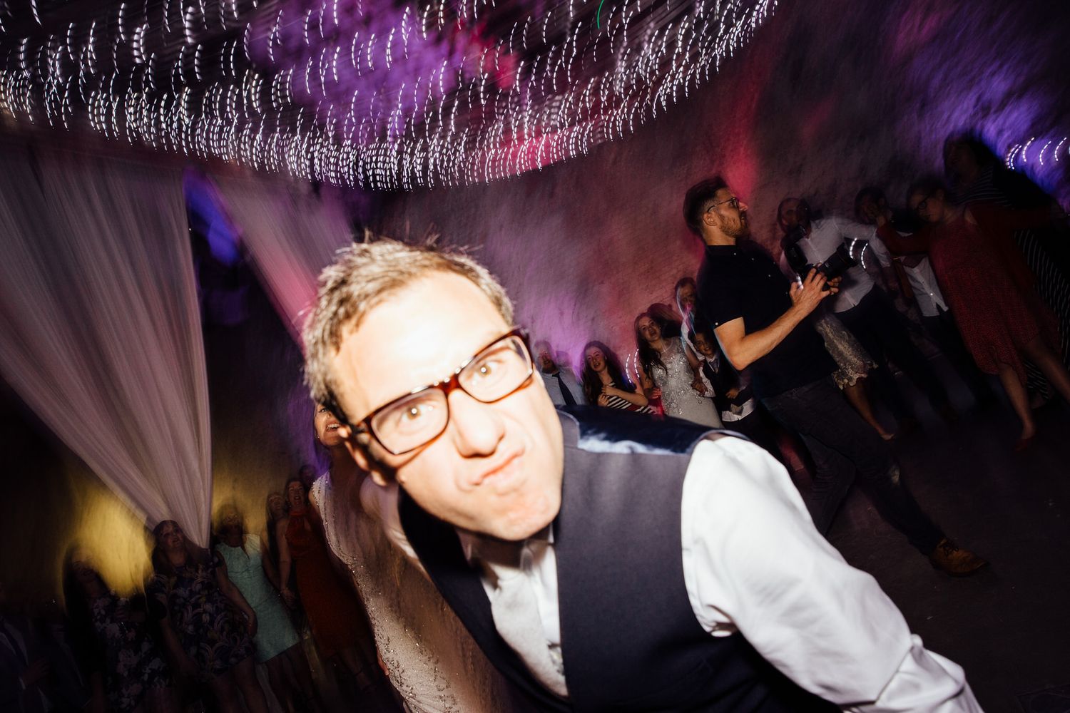 People dancing and celebrating at a nightclub with purple lighting and decorative string lights overhead.