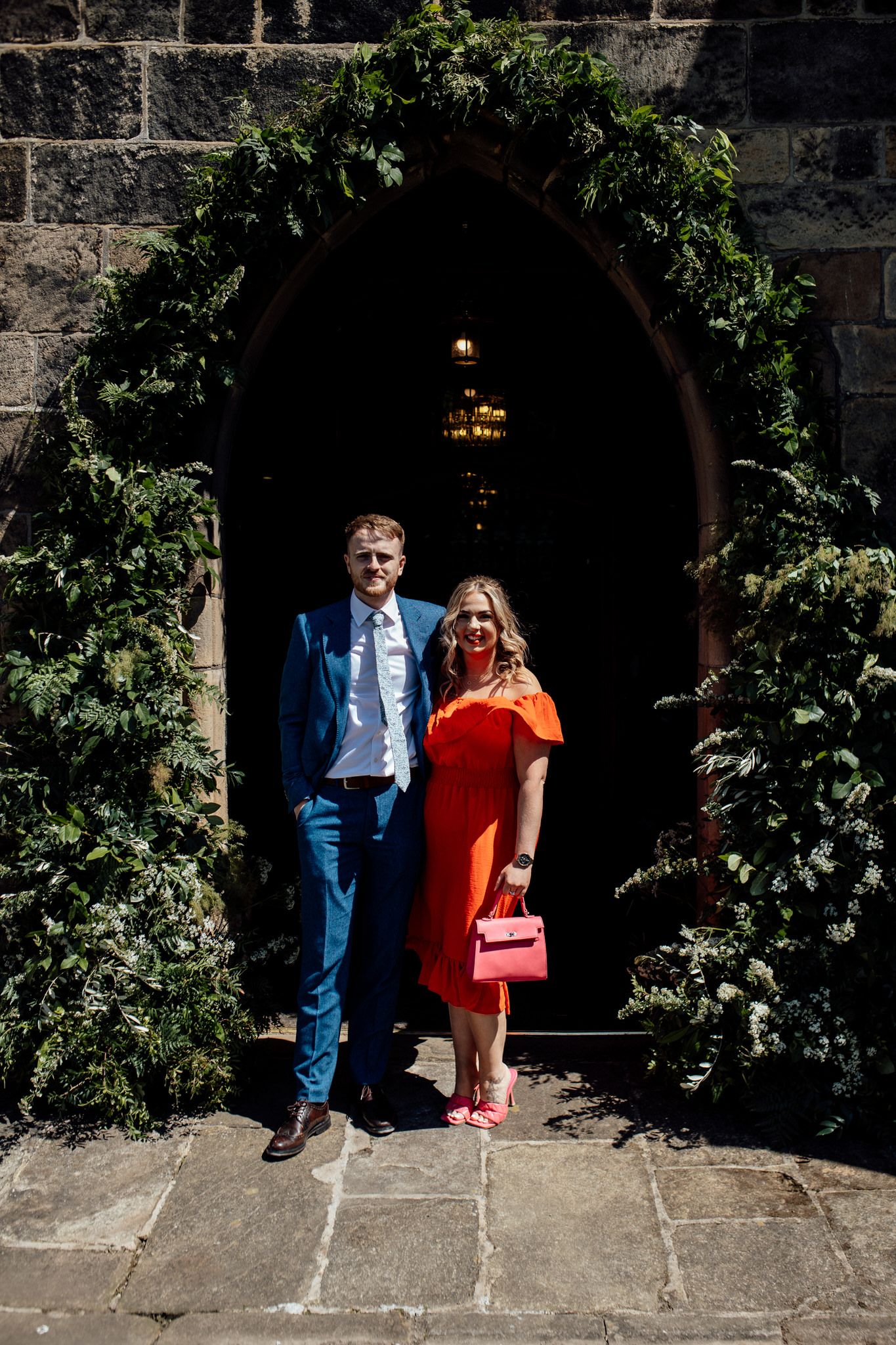 Couple standing under ivy-covered stone archway on sunny day.