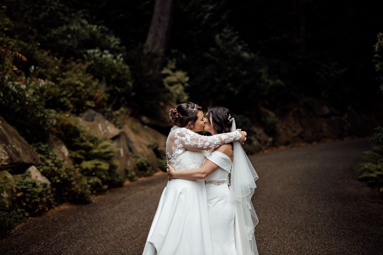 Two brides share an intimate moment on a wooded path in their white wedding dresses.
