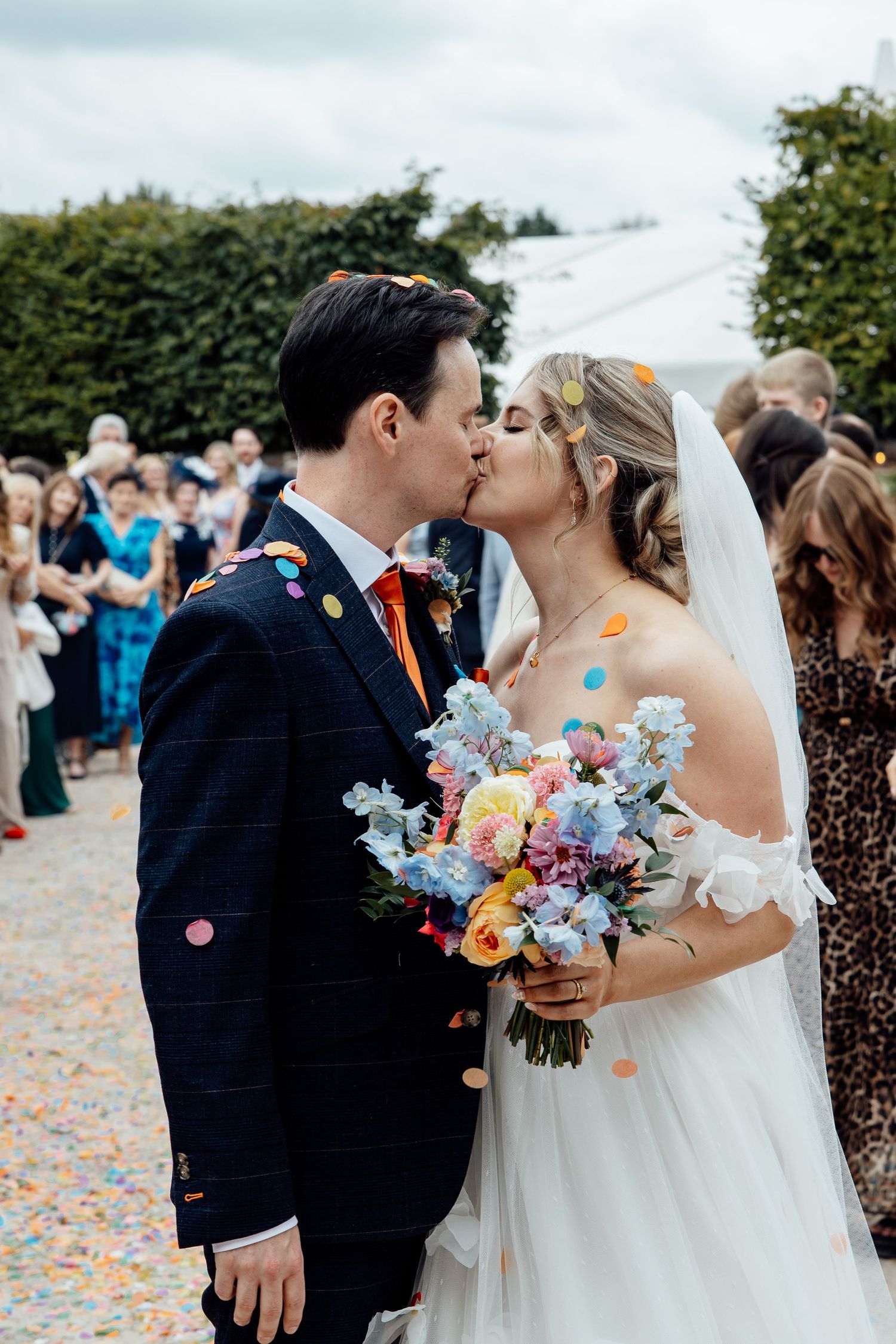 A romantic wedding kiss amid a shower of colorful confetti with guests in the background.