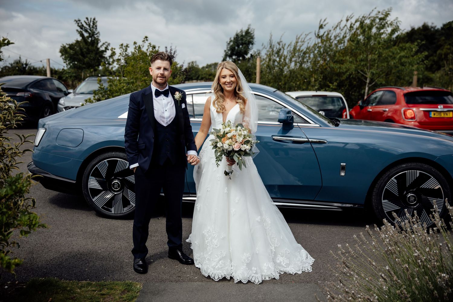 Elegant wedding party poses with luxury cars on an overcast day.