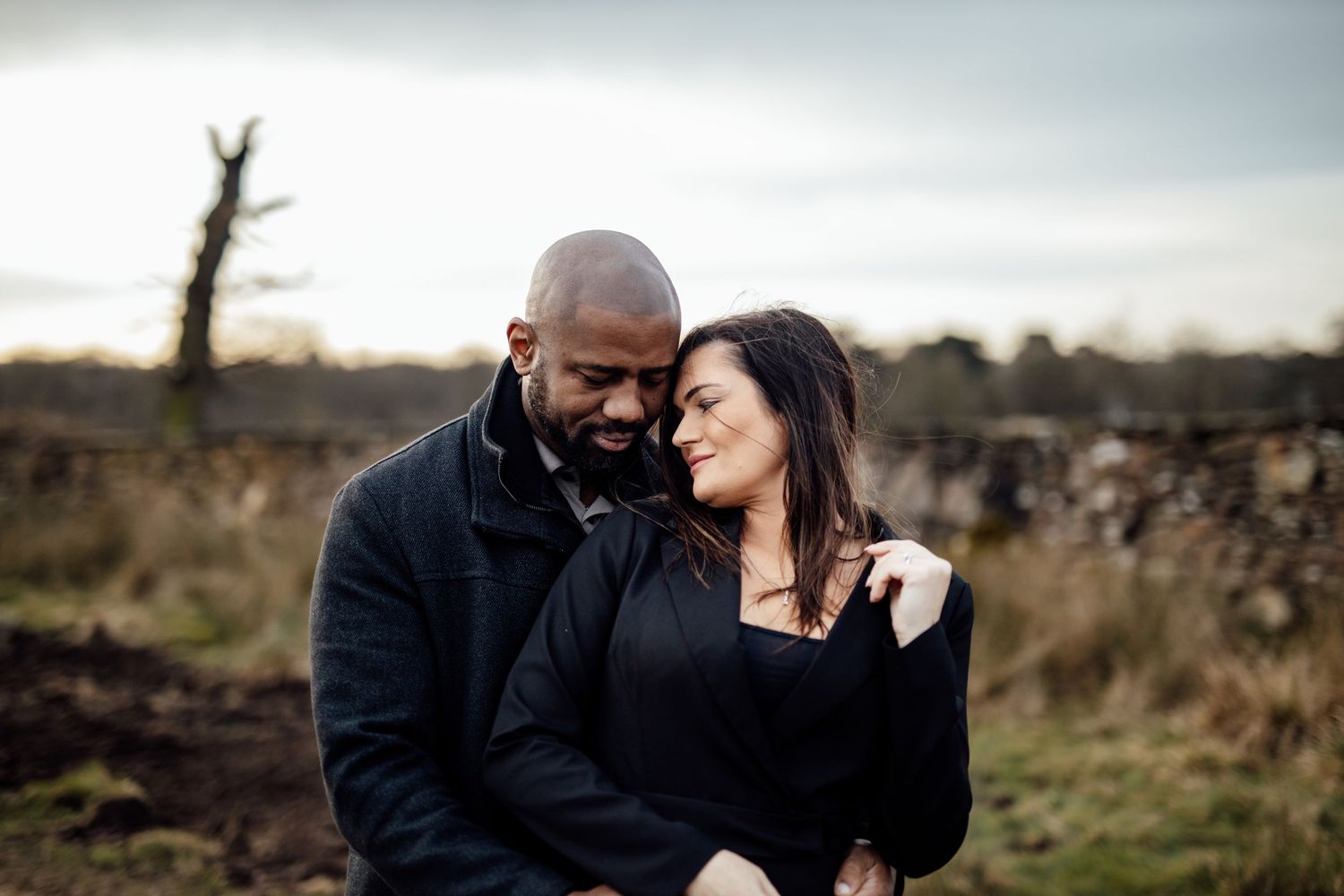 A couple shares an intimate moment together outdoors in a moody natural setting with overcast skies.