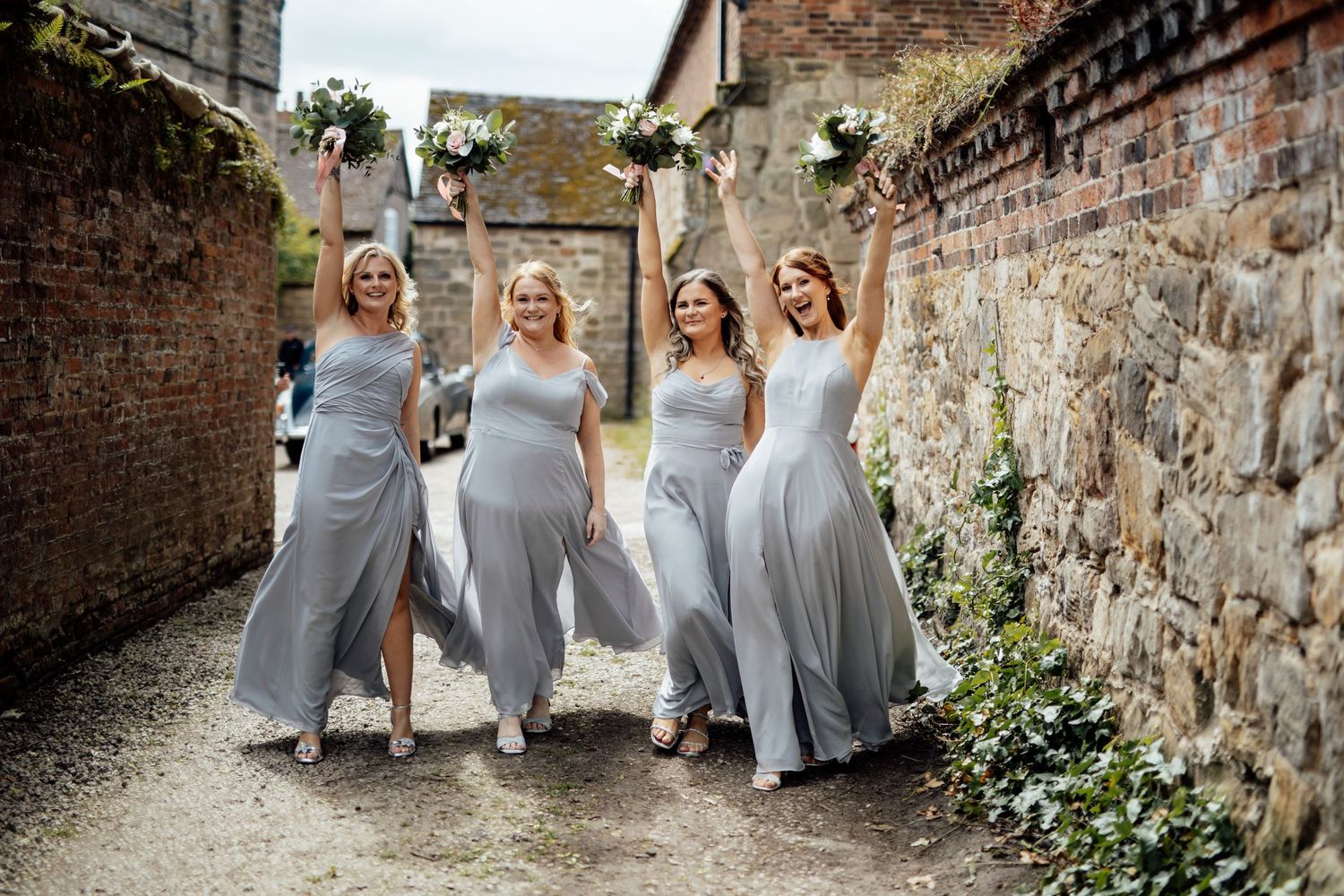 Bridesmaids in light gray dresses pose against a rustic stone wall.