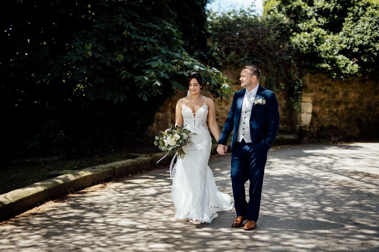 Wedding couple and guests walking along a sunny stone pathway surrounded by greenery.