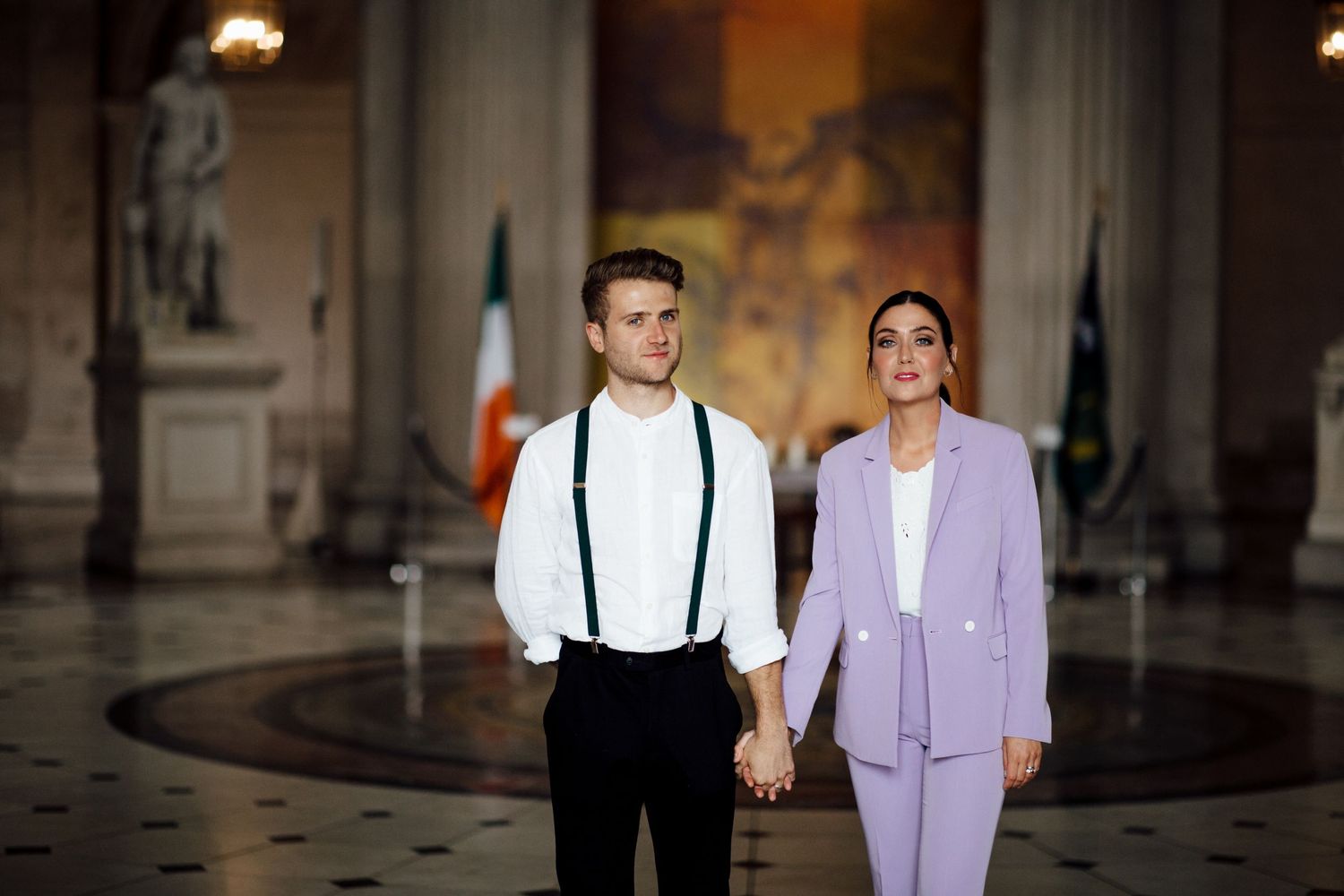 A stylish couple poses in formal wear inside a historic building with ornate details.