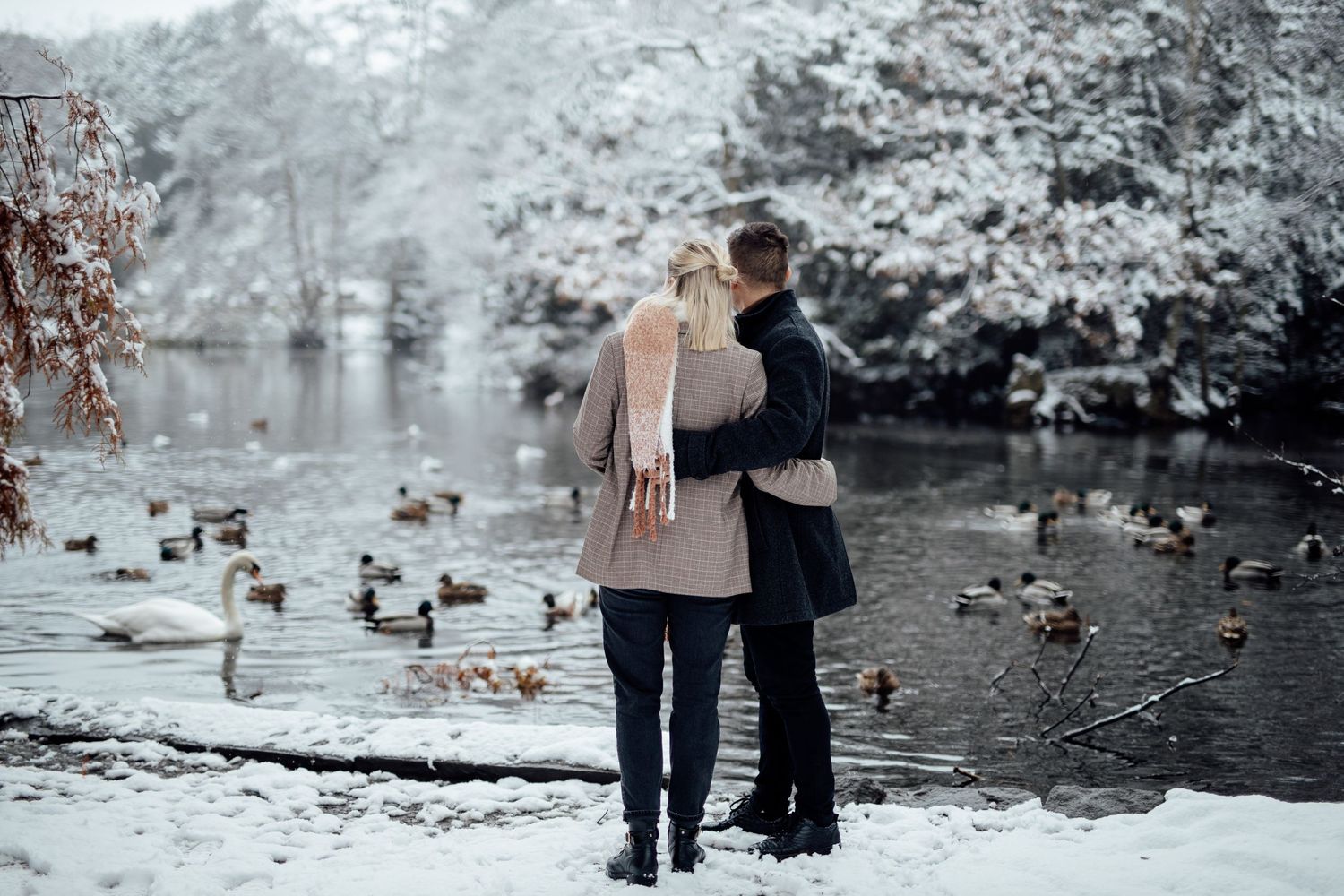 A couple shares an intimate moment by a serene pond surrounded by snow-covered trees.