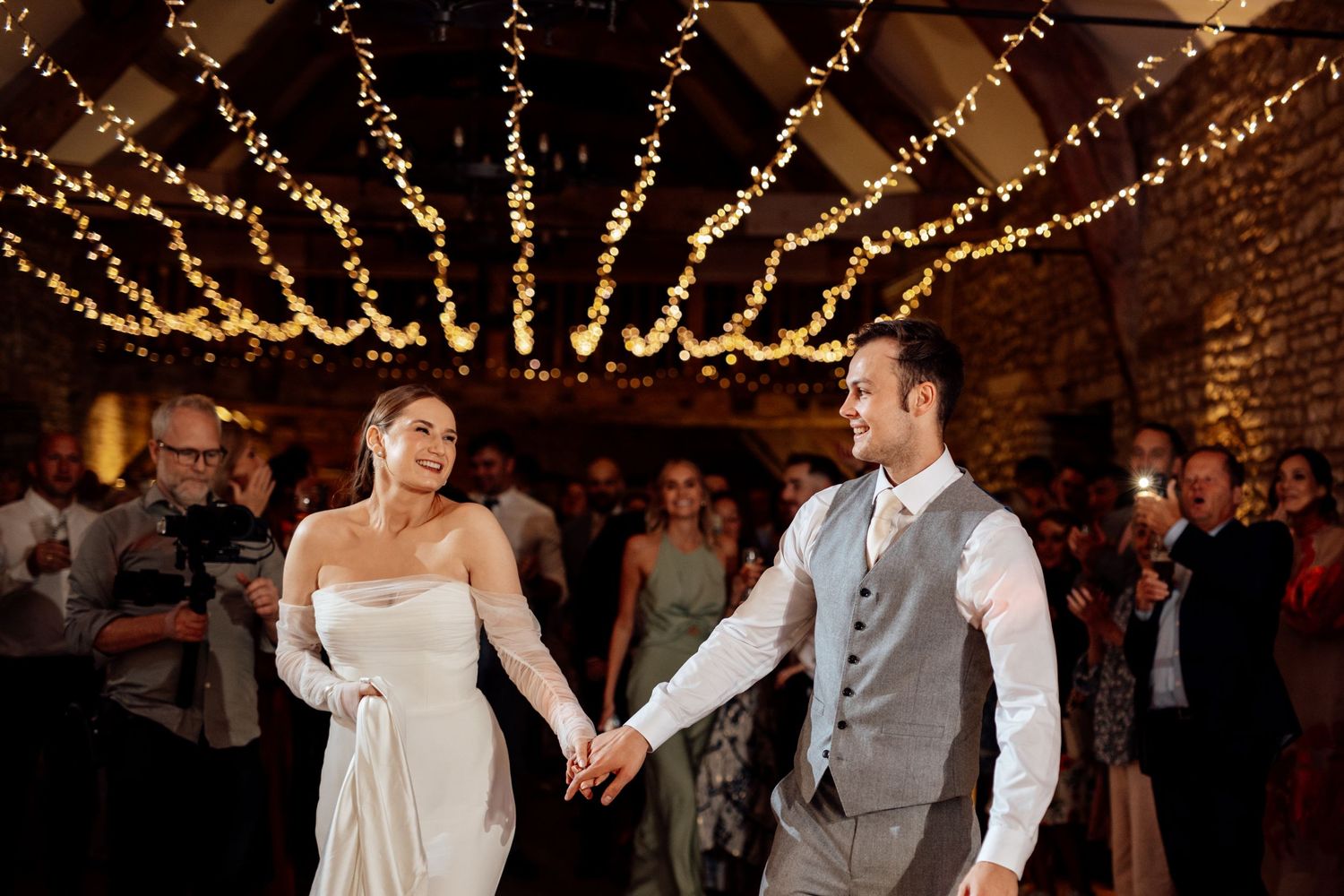 Couple dancing beneath twinkling string lights in rustic brick venue.