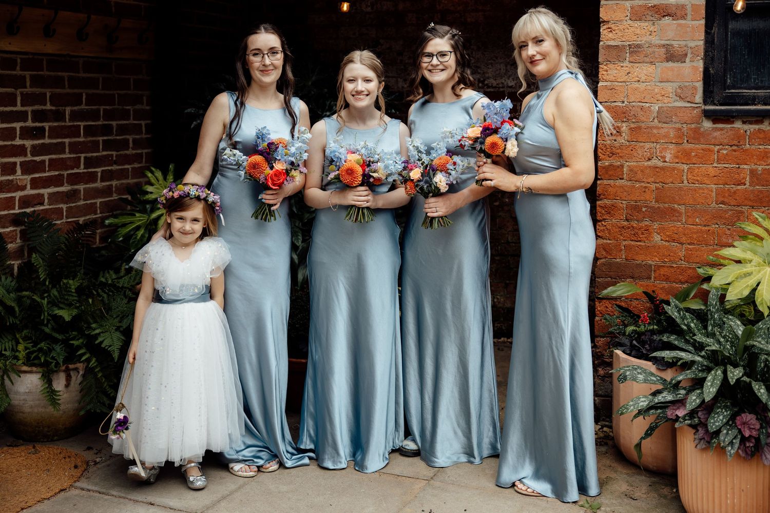 Wedding party in light blue dresses poses with bouquets against brick wall.