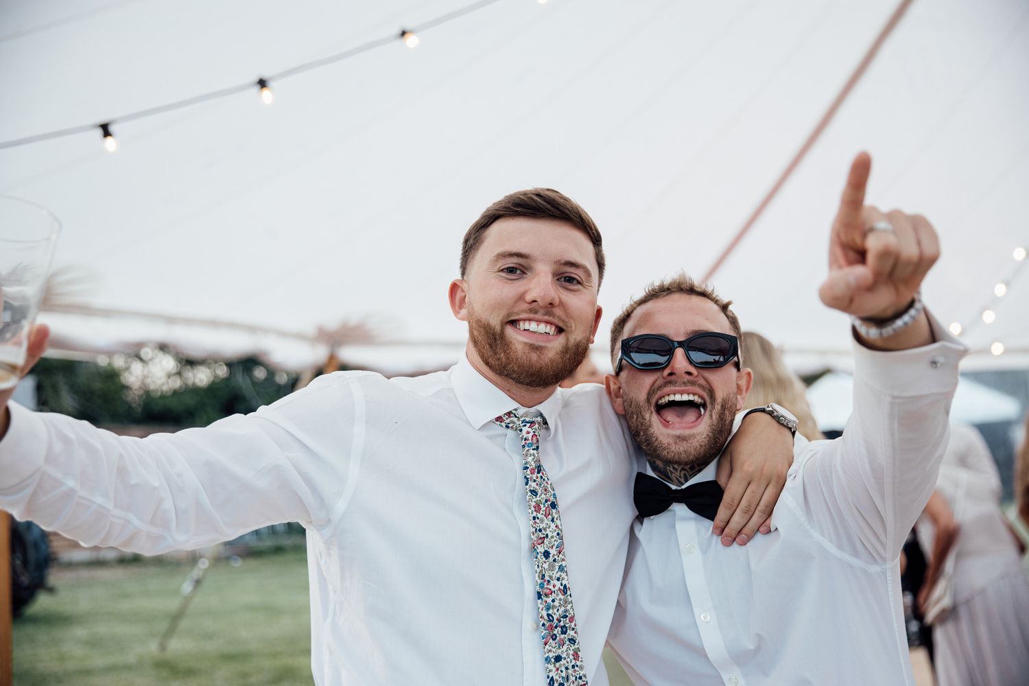 Groomsmen celebrate together outdoors during a wedding celebration with string lights overhead.