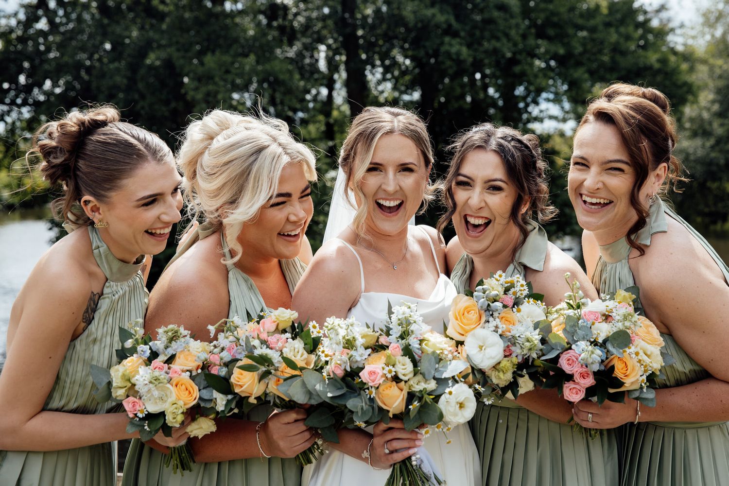Bridal party in sage green dresses laughing together outdoors during photo session.