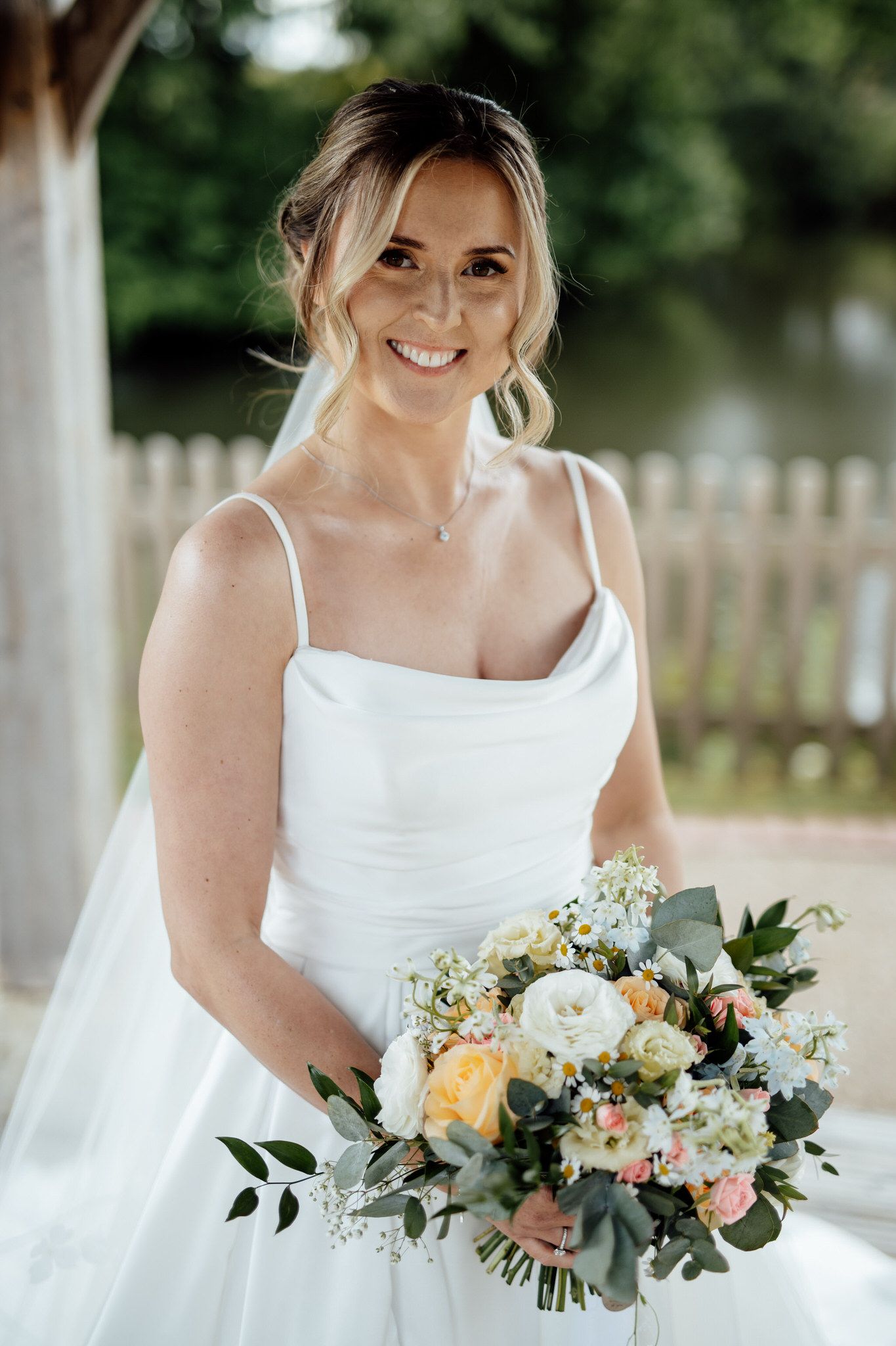 Bride in white dress holds beautiful peach and white floral bouquet with greenery.