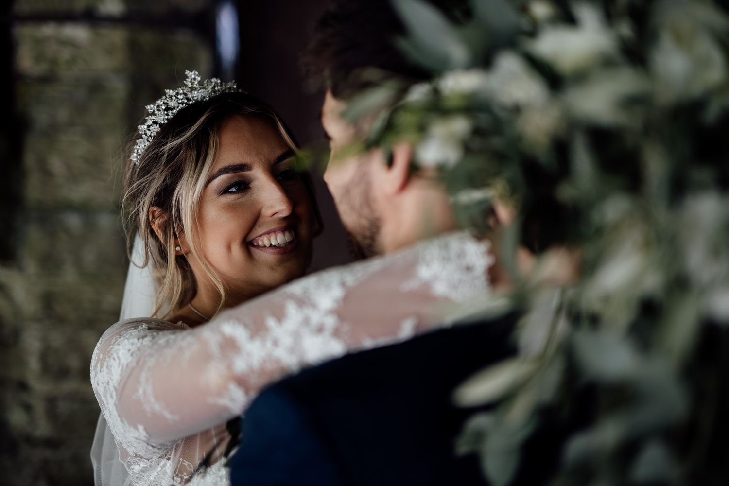 Joyful wedding portrait through foliage with bride wearing lace gown and tiara.