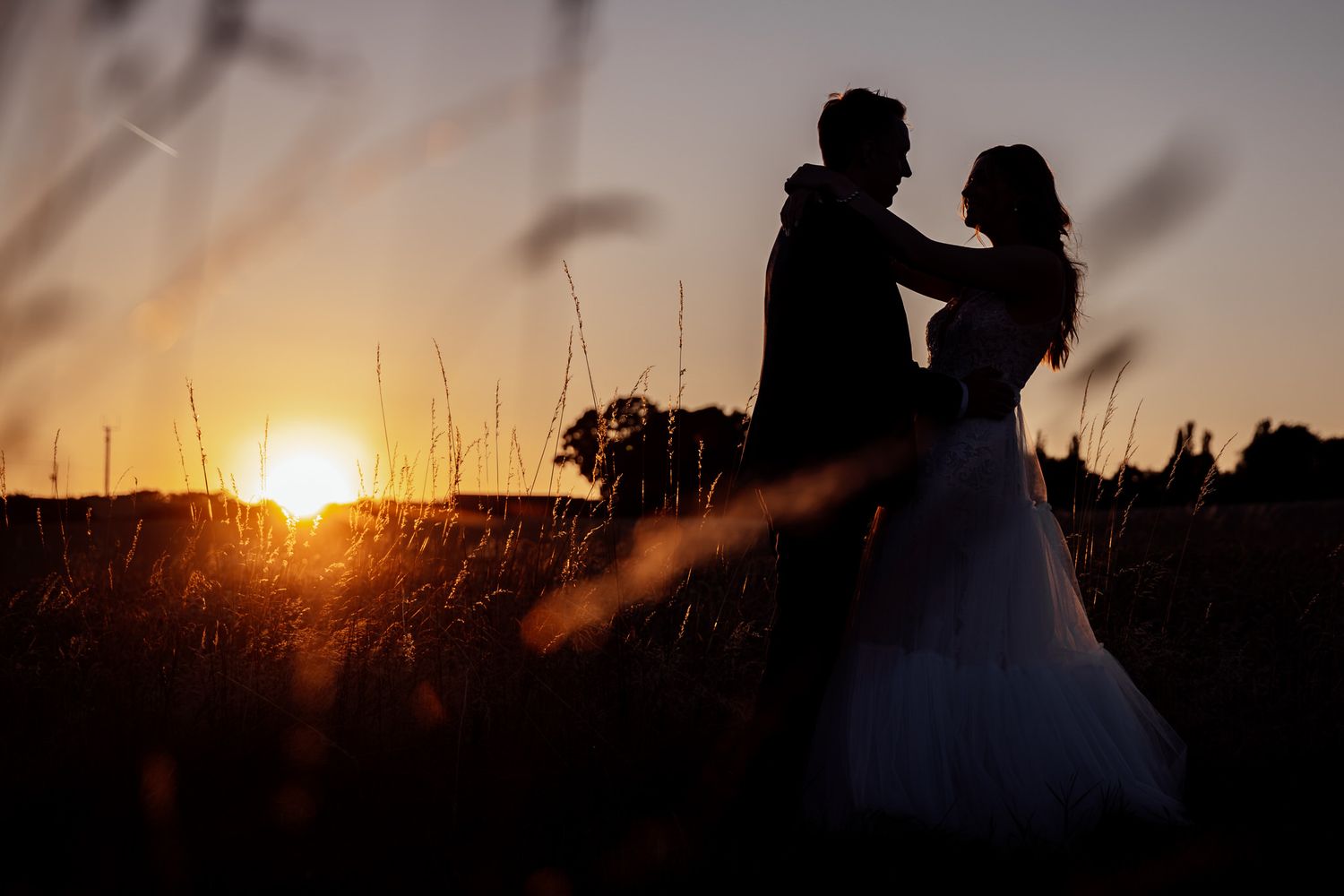Silhouettes of newlyweds against dramatic sunset in field of tall grass.