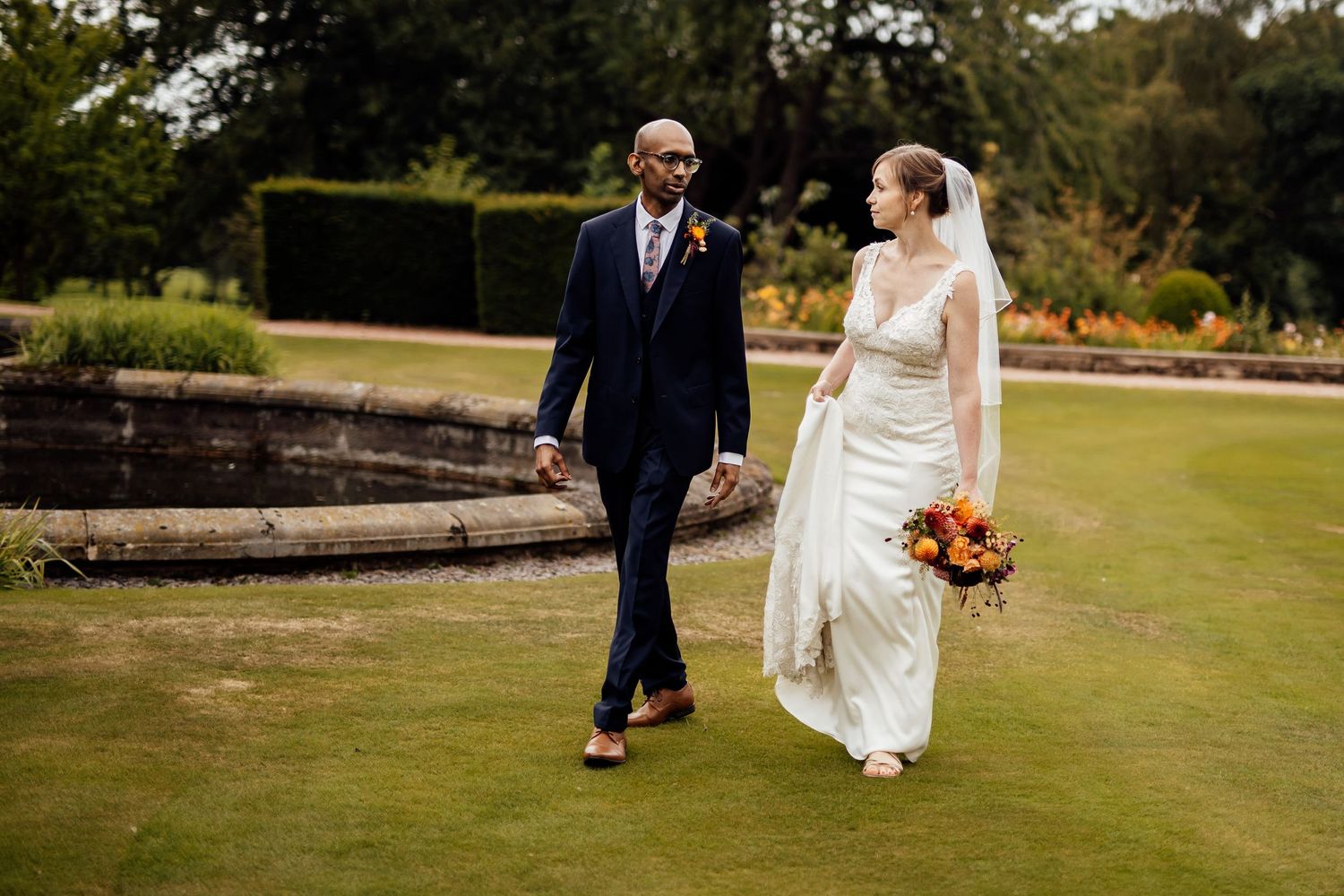 A newlywed couple walks together on manicured garden grounds with autumn-colored bouquet.