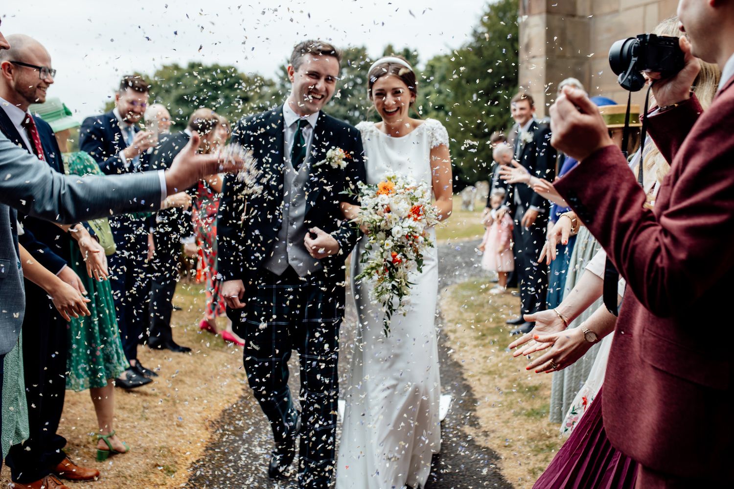 Joyful wedding celebration with guests throwing confetti in natural outdoor setting.