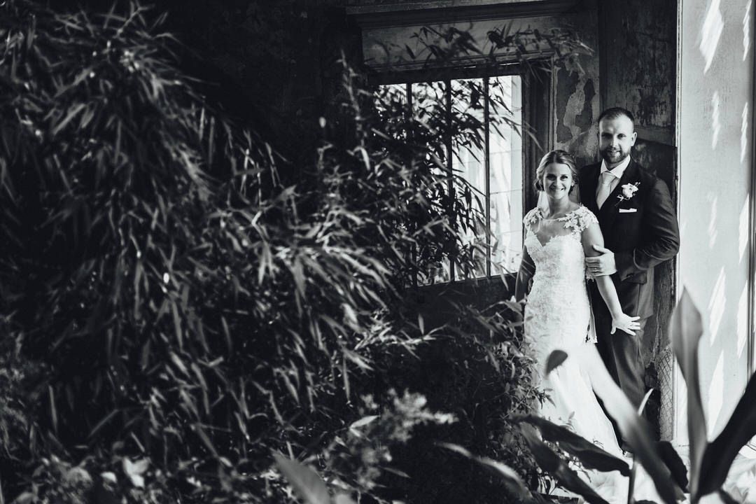 Wedding couple poses in front of lush greenery and white pillars.