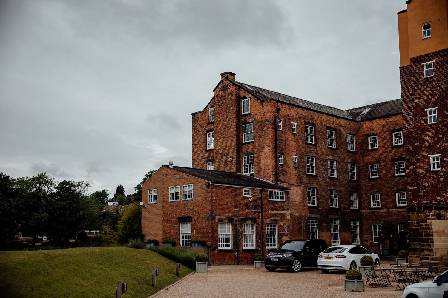 Historic red brick mill building with parked cars and curved driveway under cloudy skies.