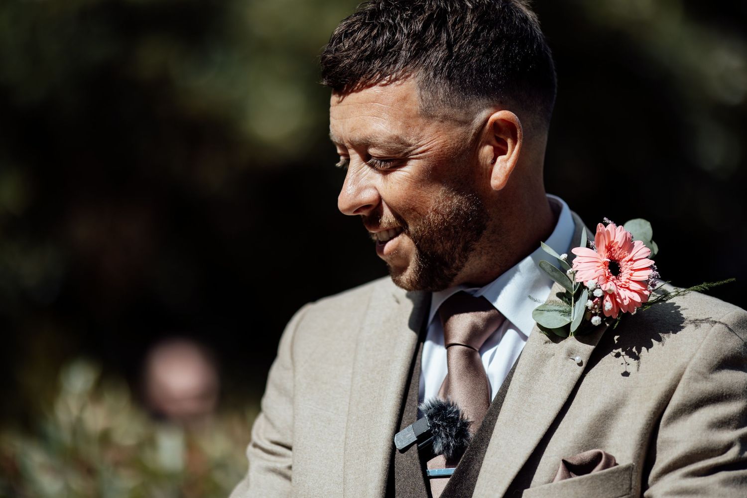 Groom in tan suit with pink floral boutonniere smiling softly outdoors on wedding day.
