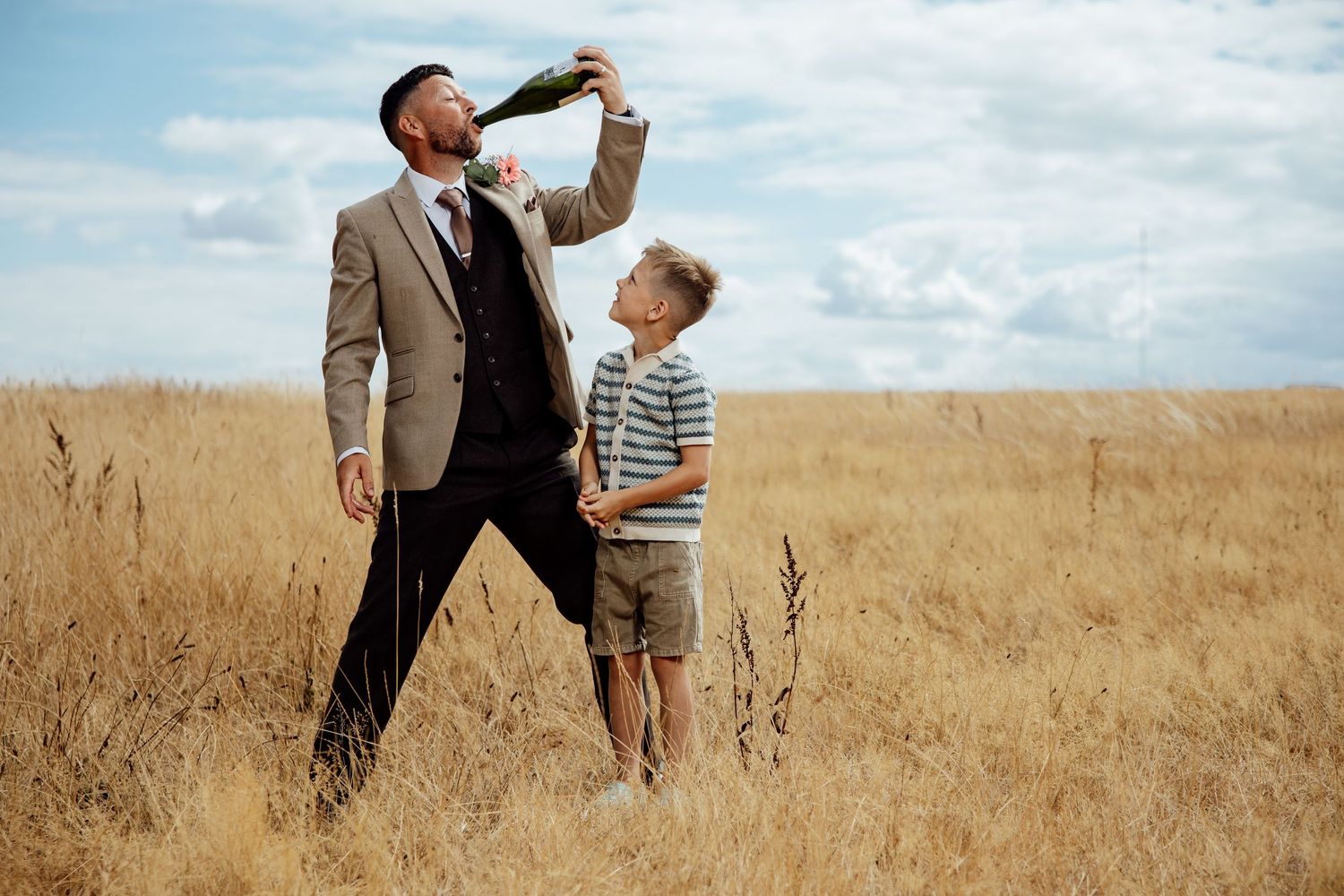 Groom drinks champagne from bottle in golden field with young boy at wedding.