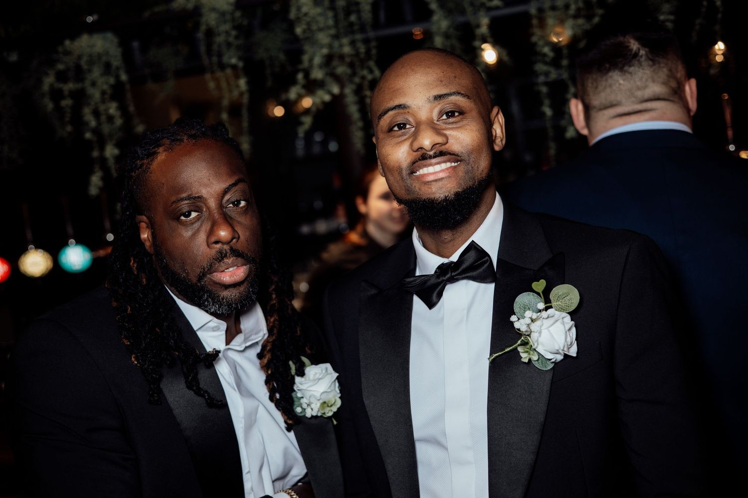 Two well-dressed men in black tuxedos with white boutonnieres smile at a wedding celebration.