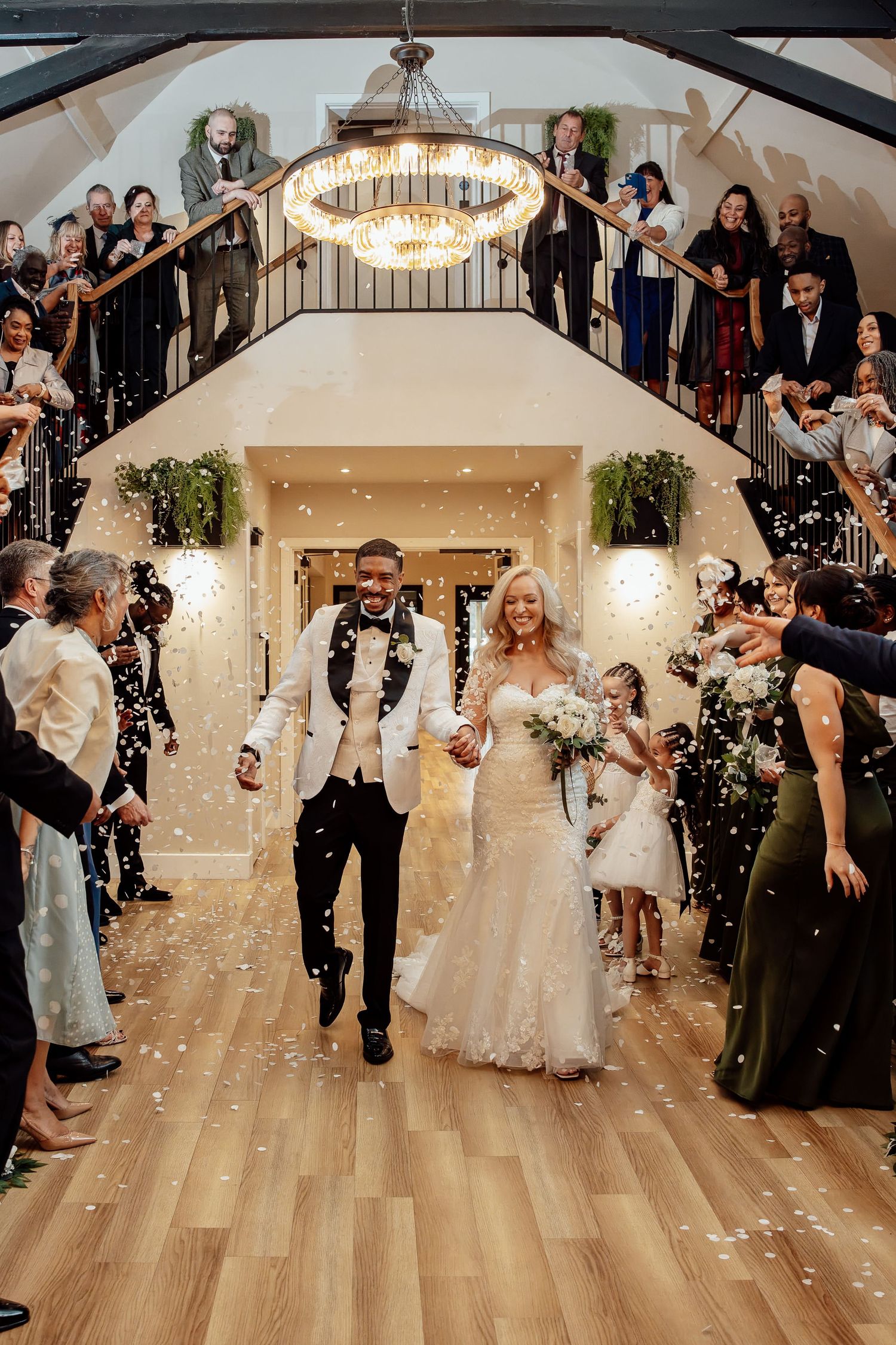 Bride and groom walk through confetti shower at elegant wedding venue with grand staircase and chandelier.