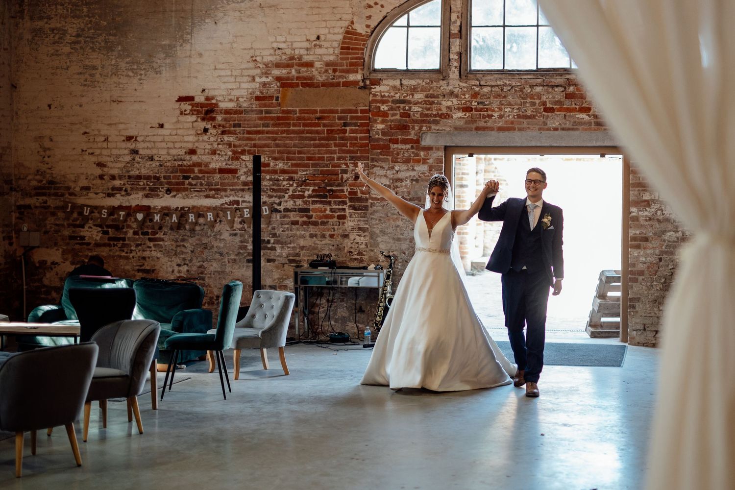 A couple dances in a rustic brick-walled venue with vintage teal chairs and arched windows.