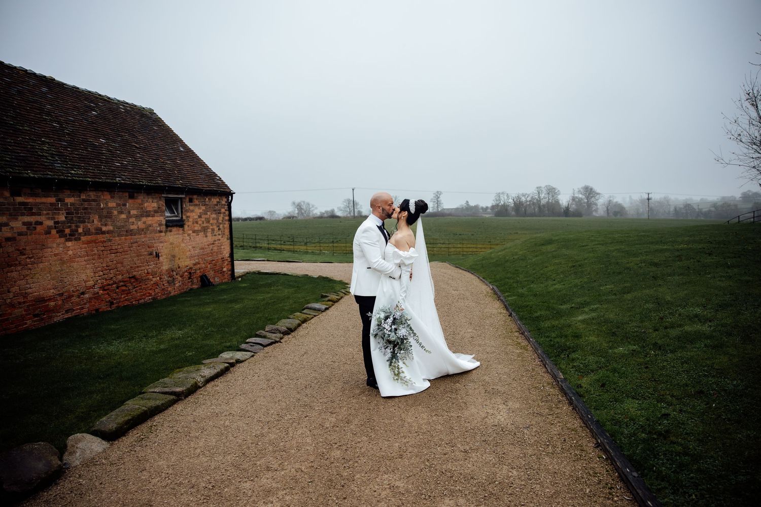Wedding couple embracing on path near rustic barn in misty countryside setting.