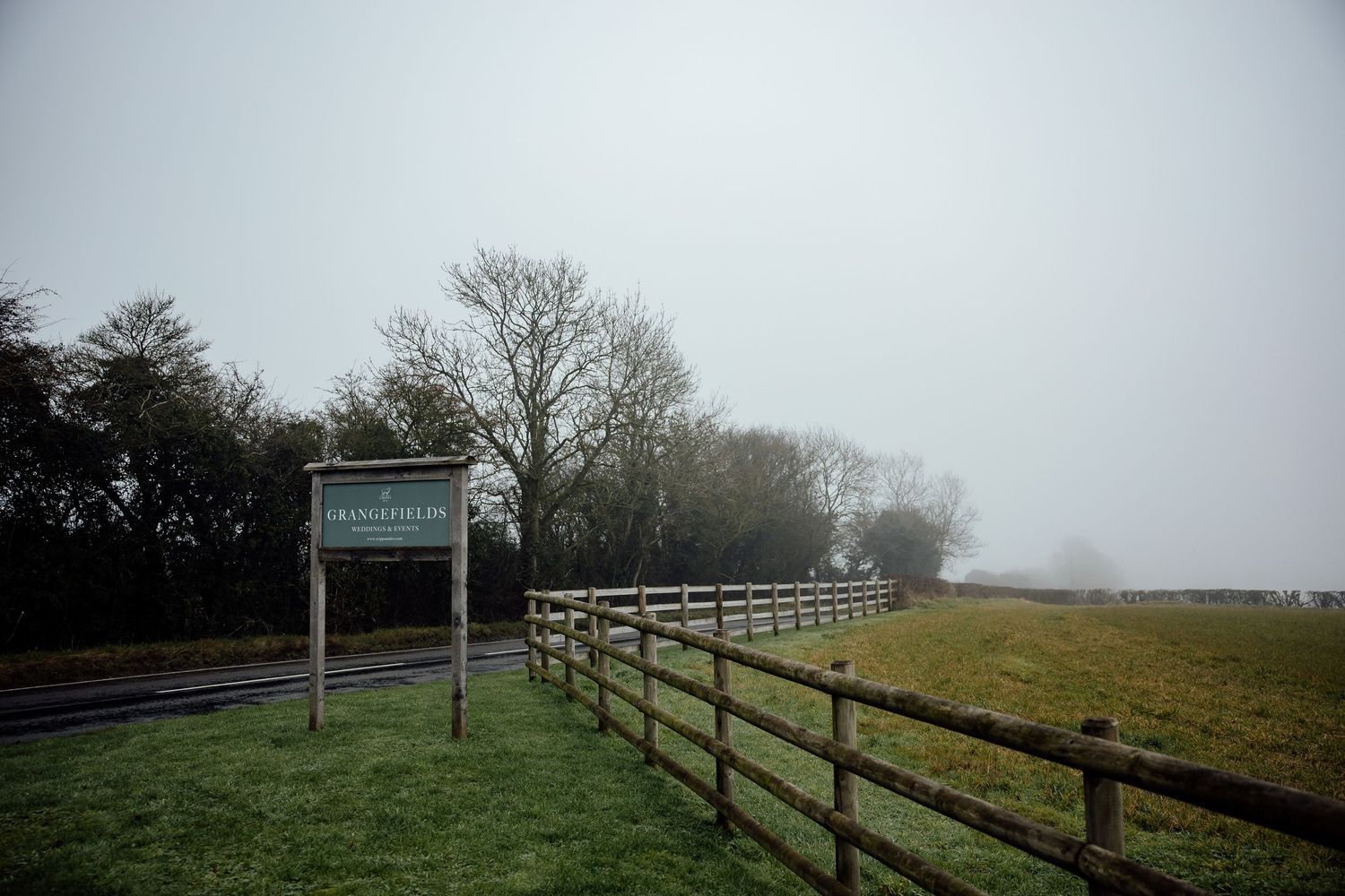 A wooden fence stretches along a misty countryside field with a sign post visible in the foreground.