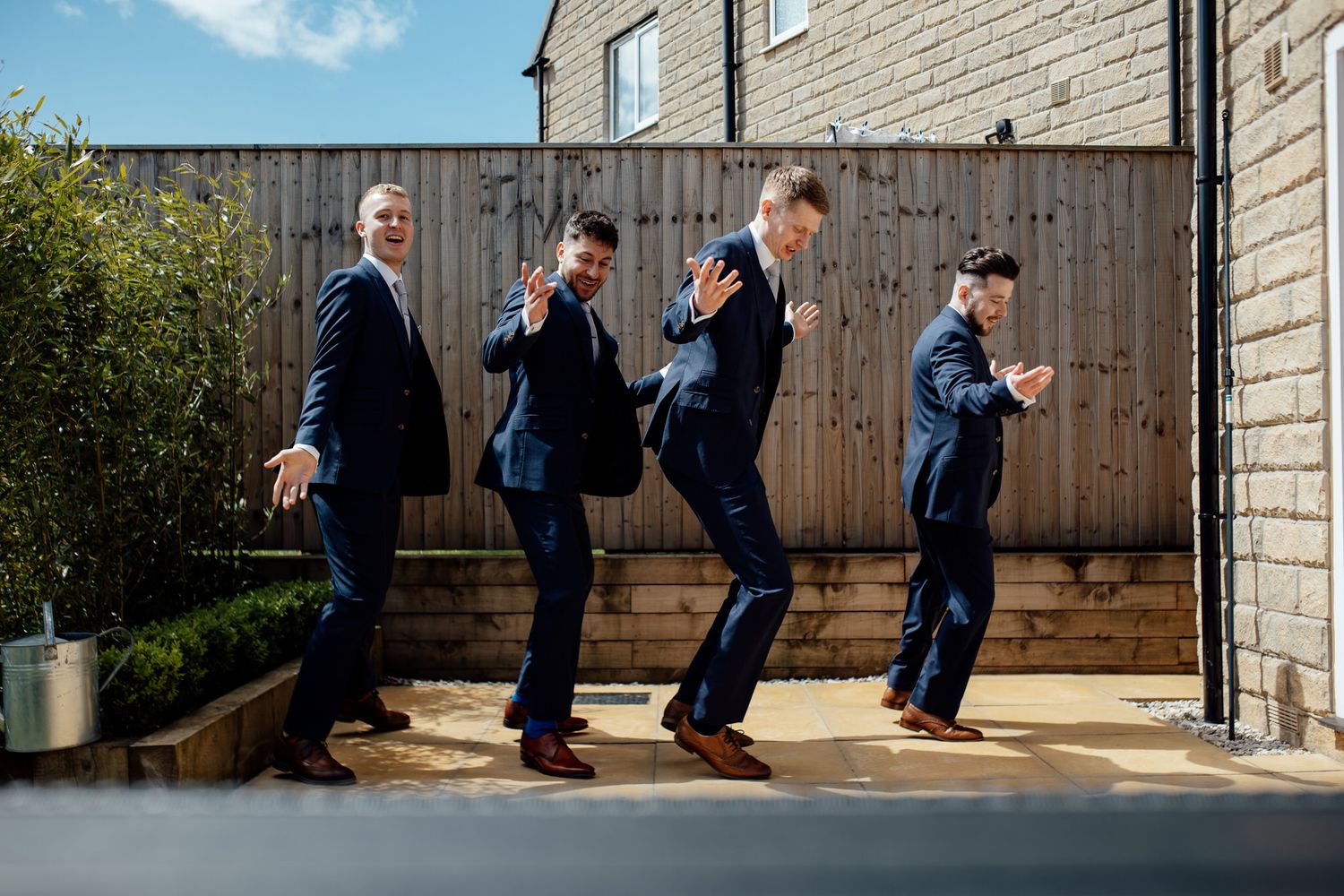 Fun sequence of groomsmen jumping in navy suits against wooden fence backdrop.
