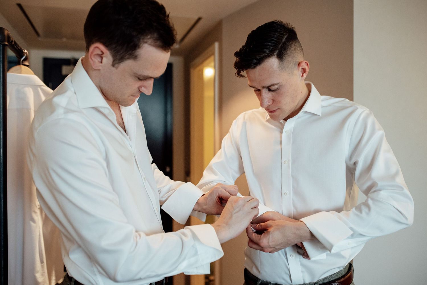 Groomsmen help each other prepare and adjust formal white dress shirts before a wedding ceremony.