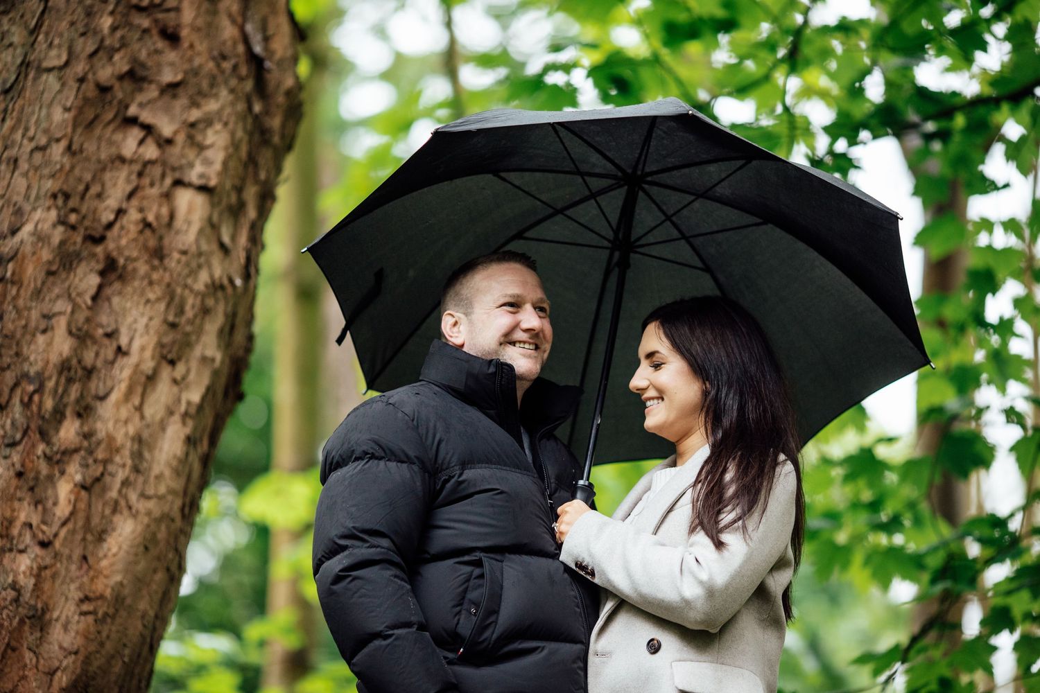 A couple shares a joyful moment under a black umbrella in a lush forest setting.