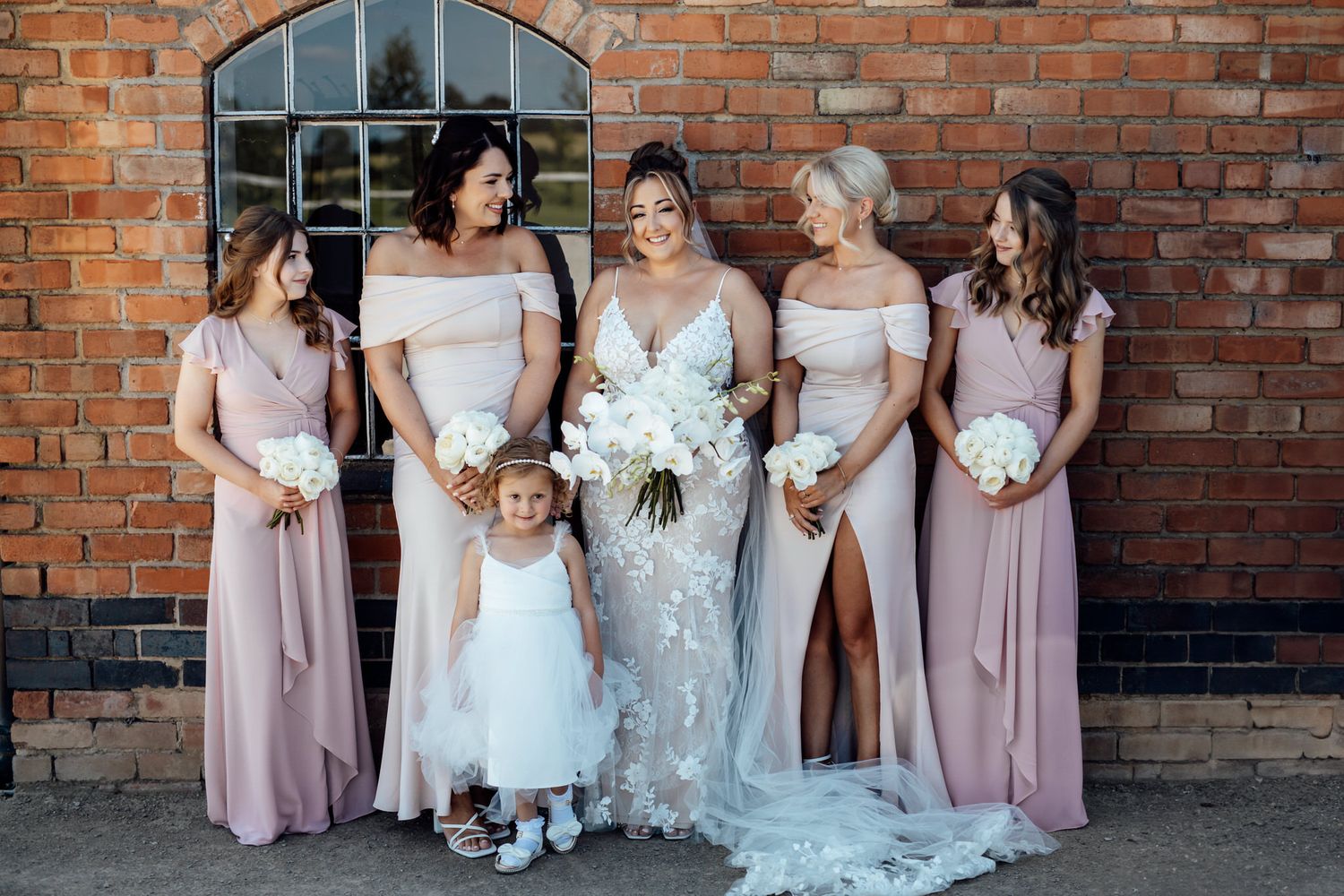 Bridal party in pink and white dresses pose against brick wall with bouquets.