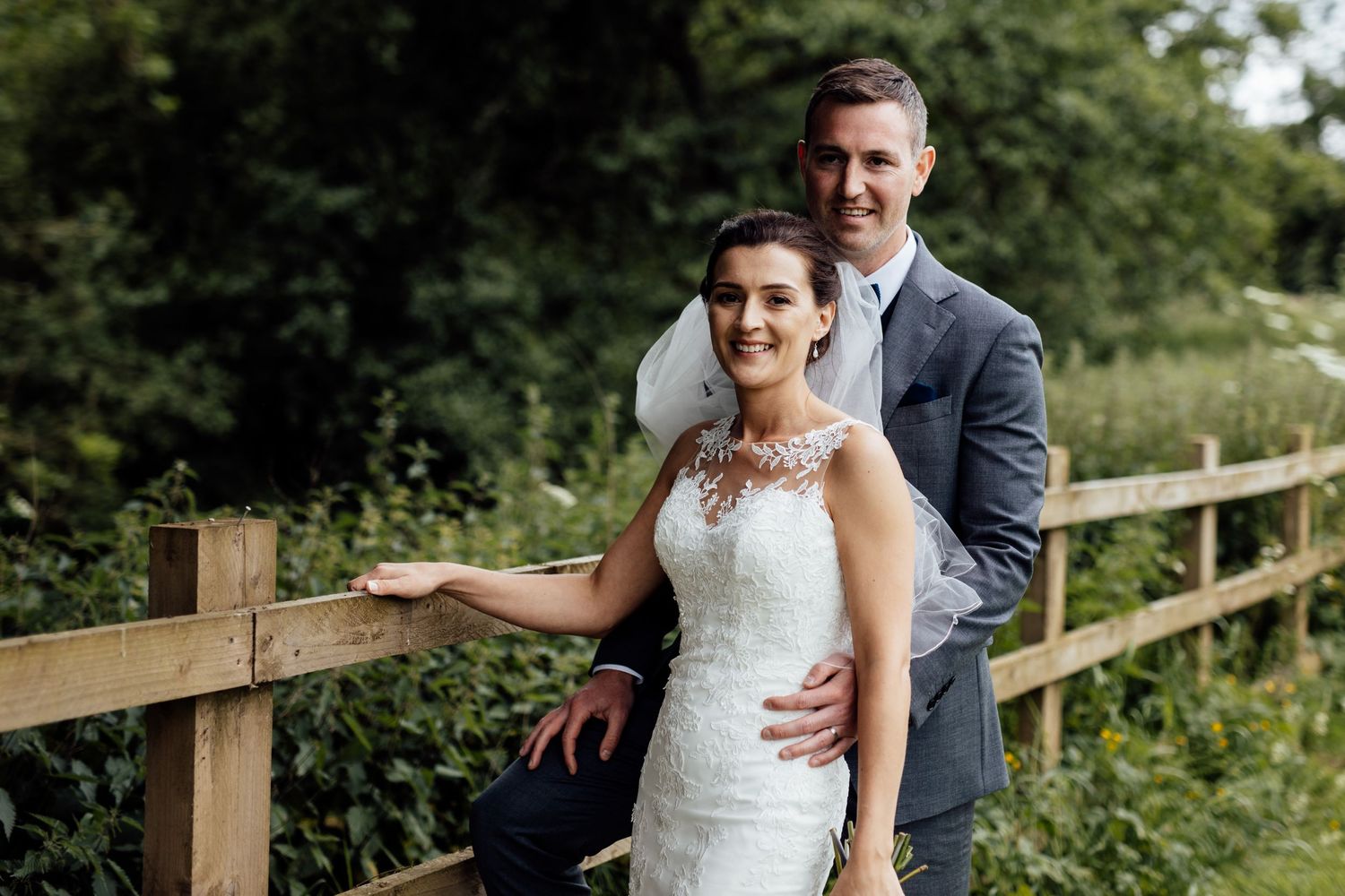 Newlyweds pose for portraits along wooden fence in outdoor garden setting.