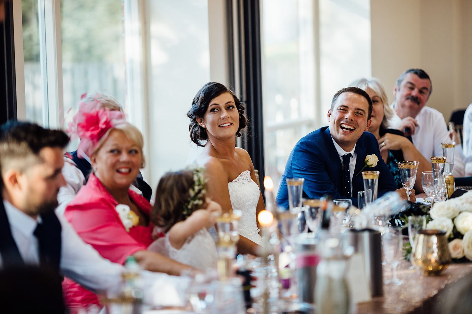 Wedding reception guests dining and socializing at tables by sunlit windows.