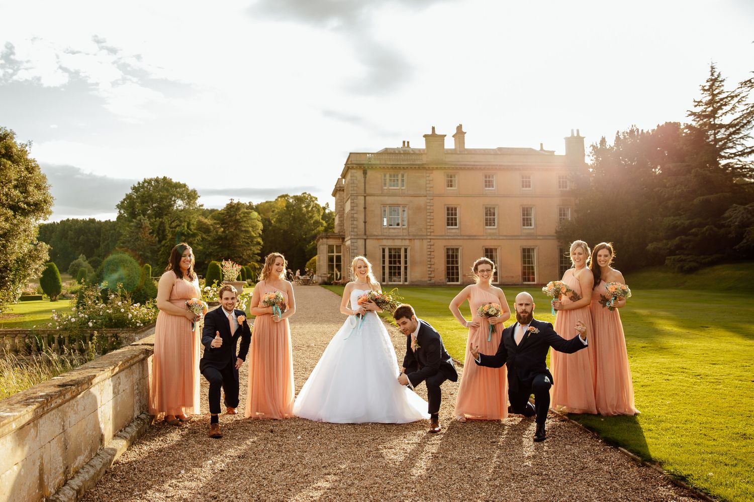Wedding party poses on grassy lawn at sunset with bridesmaids in peach dresses.