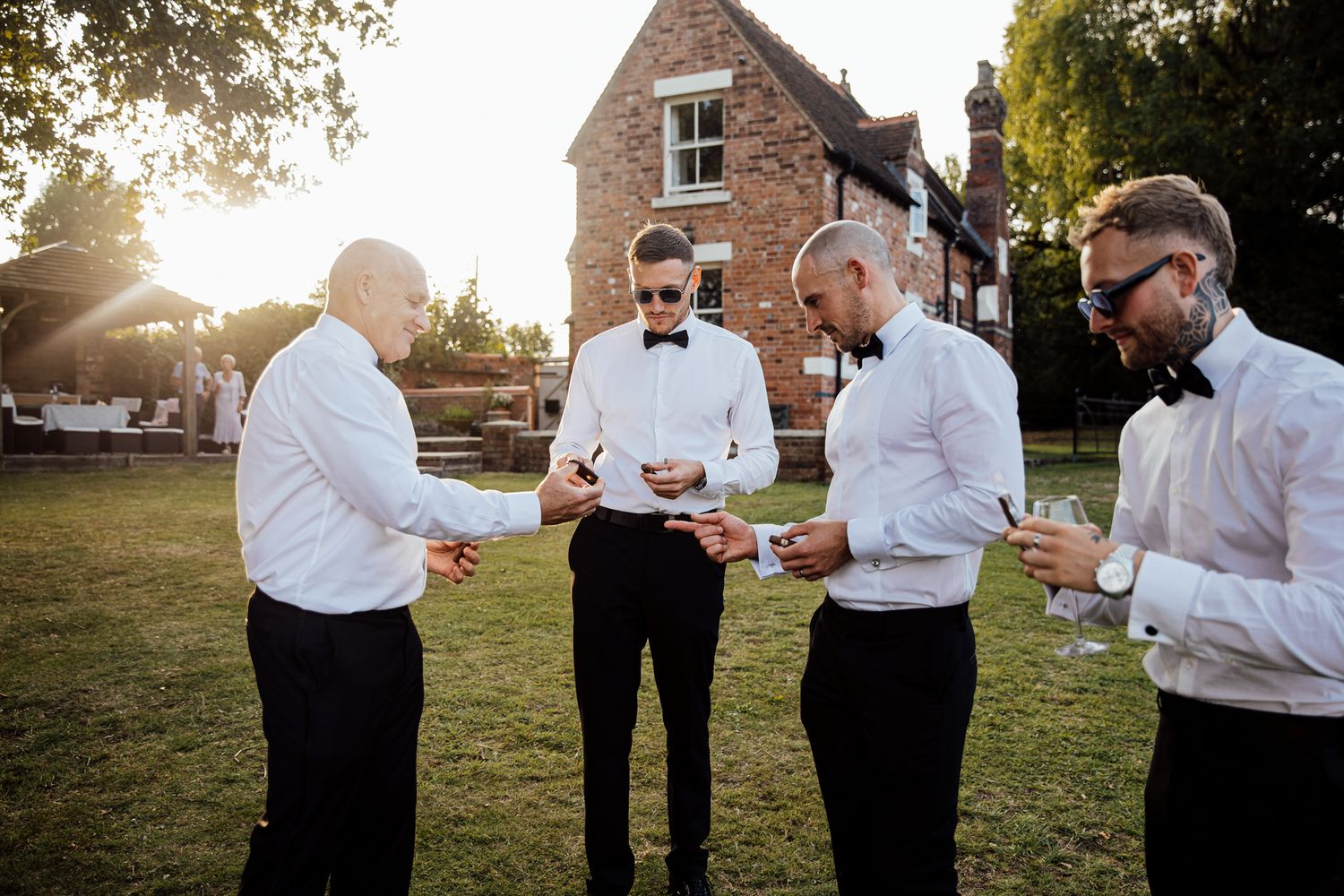Groomsmen in formal attire checking their phones while standing on grass near a brick building.