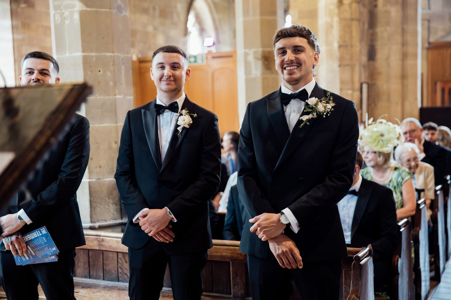 Groomsmen in black tuxedos share laughs and smiles during a wedding ceremony in a historic church.