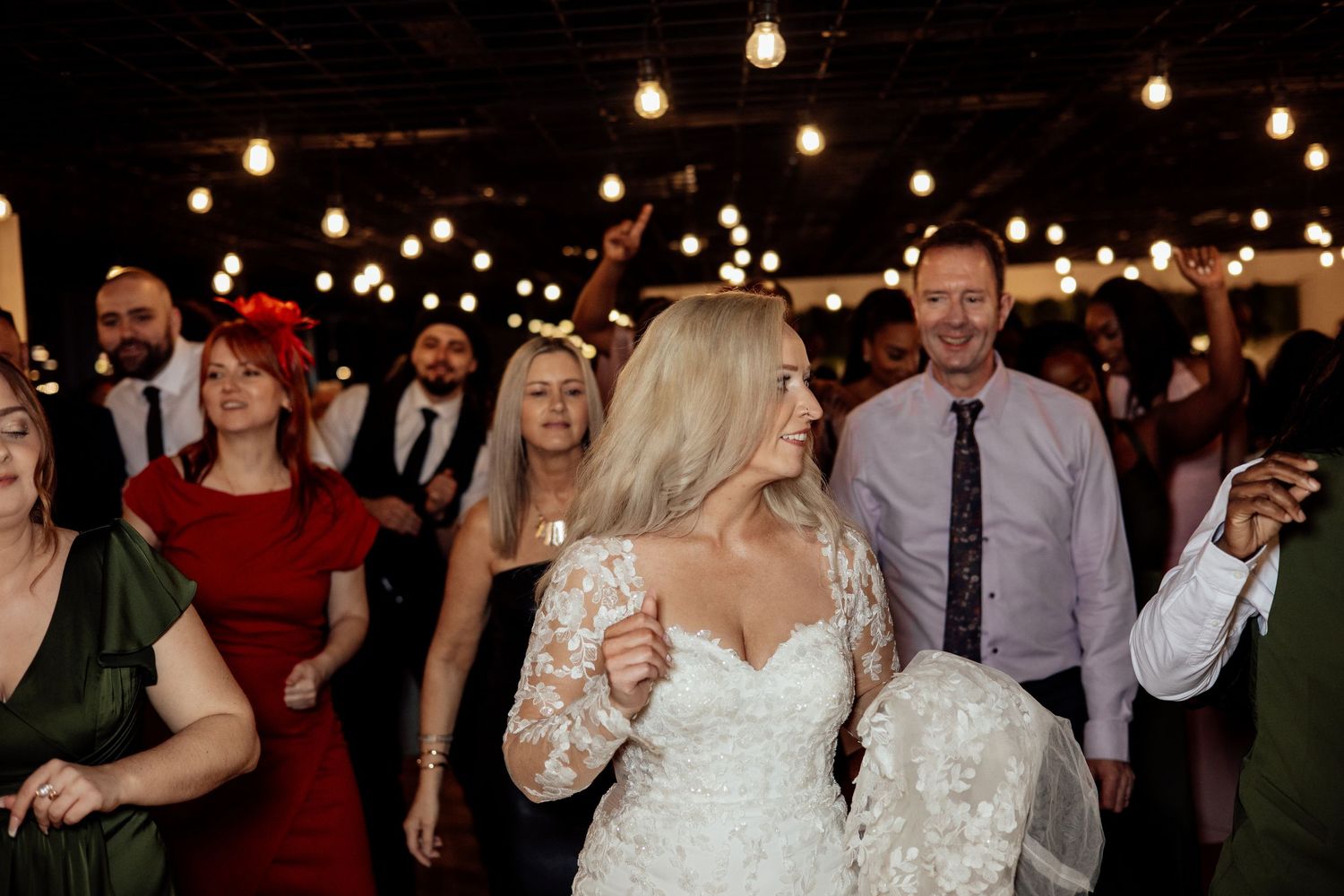 A bride dances joyfully with wedding guests under warm string lights at the reception.