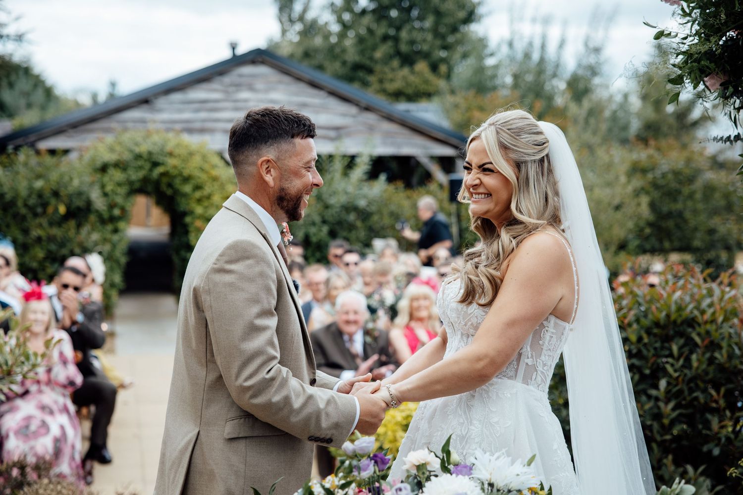 Bride and groom holding hands smiling during outdoor garden wedding ceremony with guests seated behind.