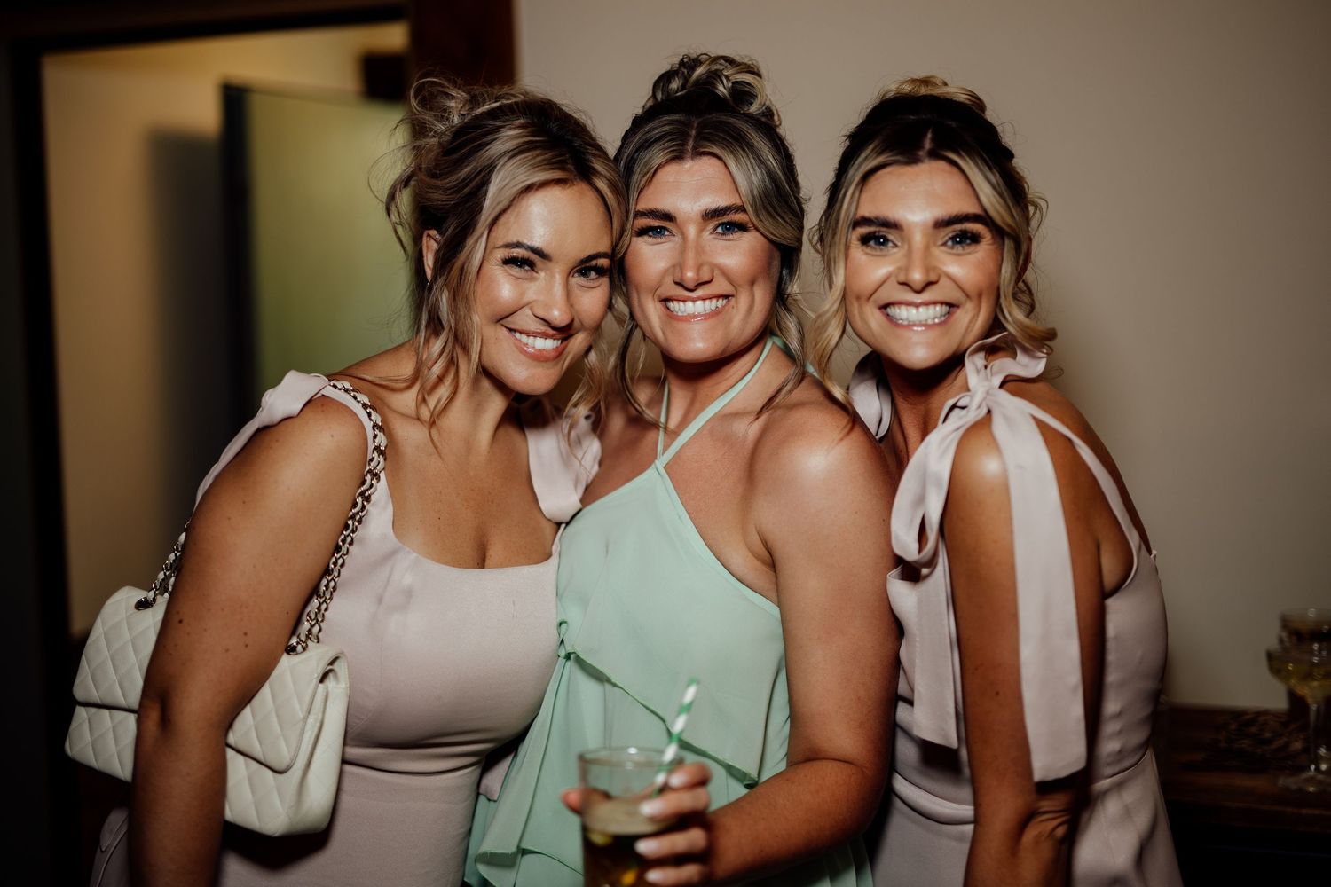 Three smiling women pose together at a wedding celebration wearing elegant dresses in neutral and mint tones.