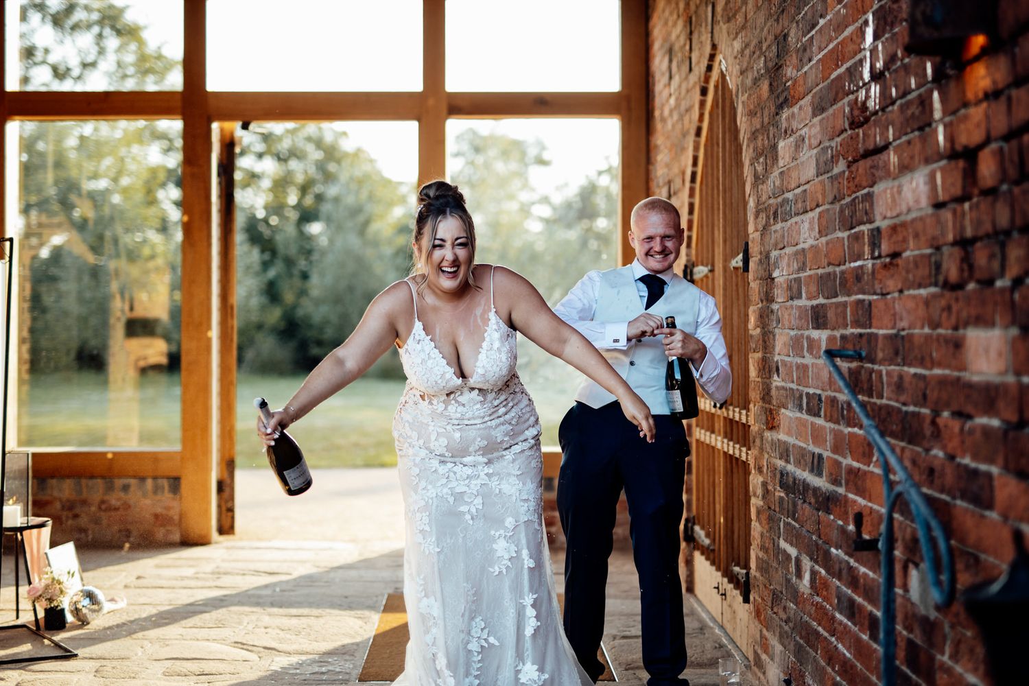 A joyful celebration moment at a rustic brick venue with a lace dress and champagne bottle.