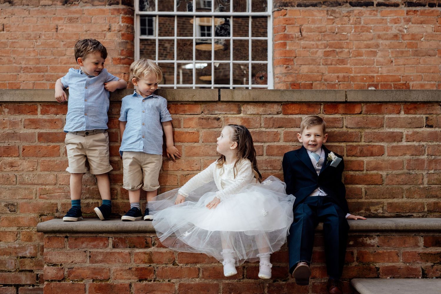 Children in formal attire sitting and standing by brick wall at wedding.