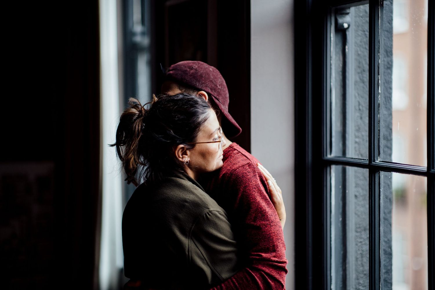 Two people embrace near a window in a dimly lit interior setting.