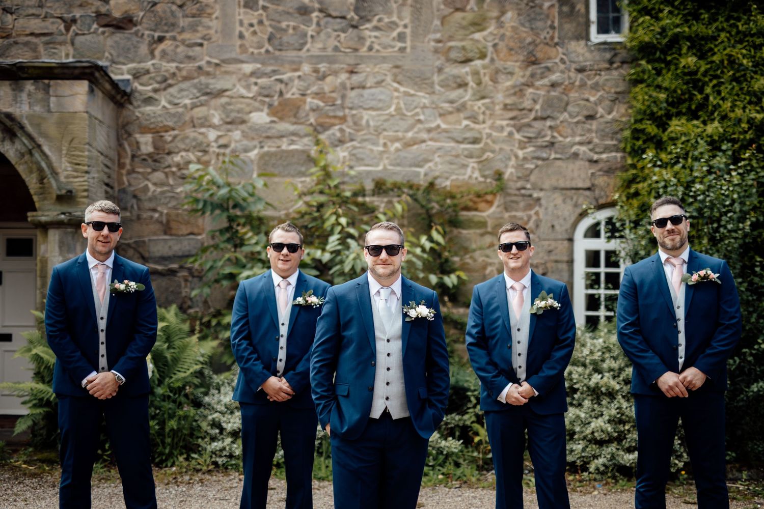 Groomsmen in navy suits and sunglasses pose together against a stone wall backdrop.